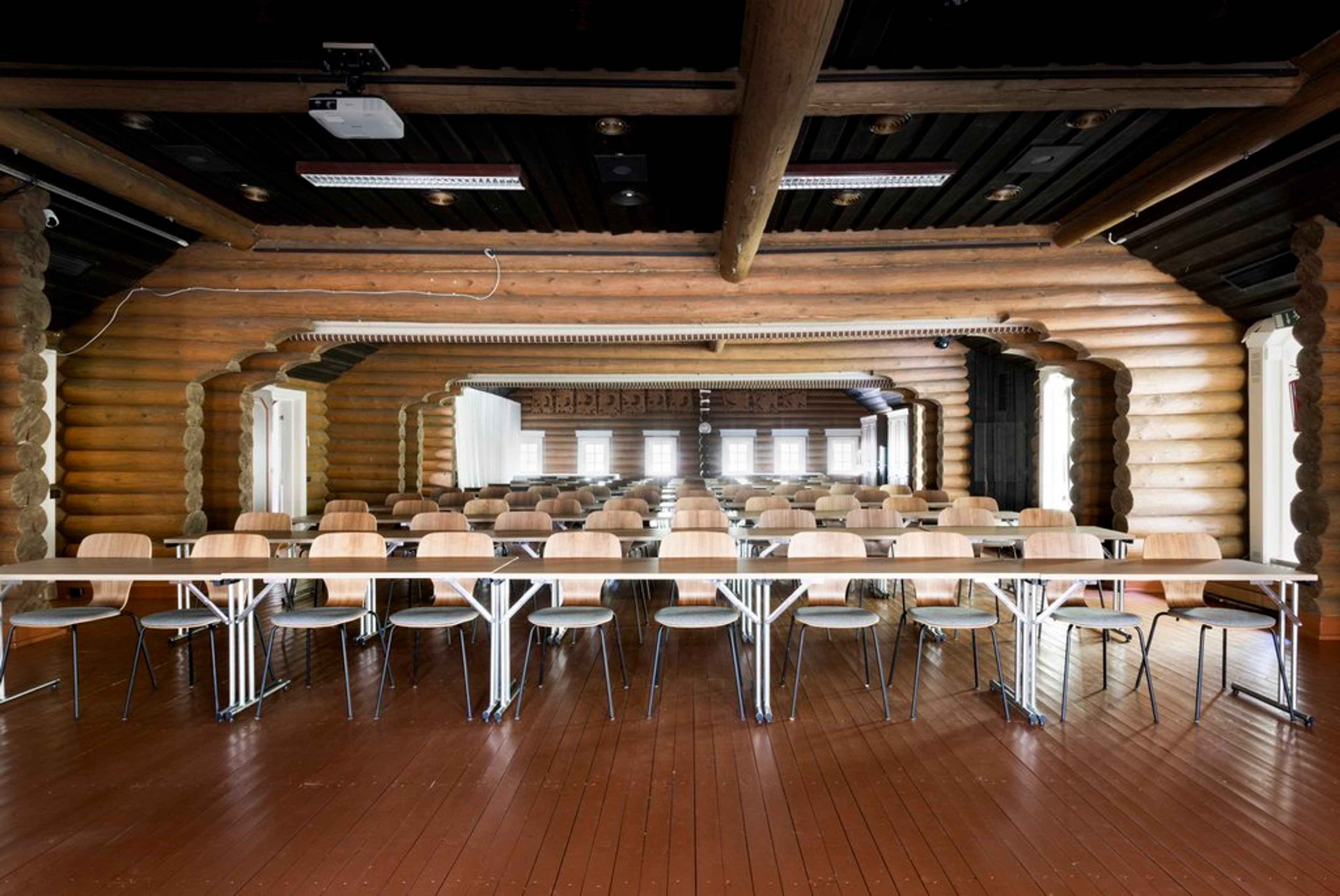 Log-walled conference room with long tables and chairs in rows. Dark ceiling with wooden beams and hardwood flooring.
