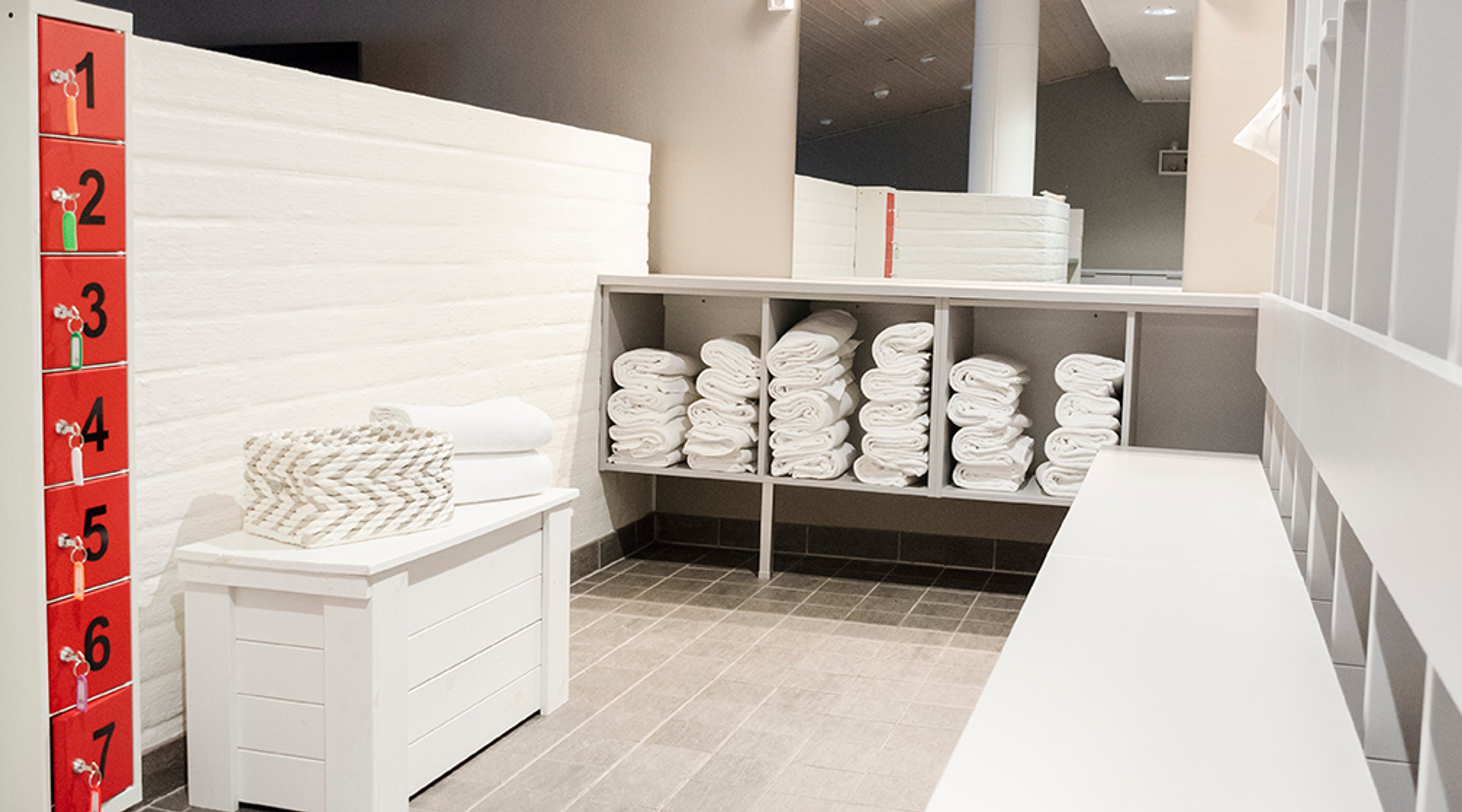 Saunan pukuhuone, jossa on punaiset numeroidut lokerot vasemmalla, valkoisia pyyhkeitä hyllyissä ja harmaa laattalattia.