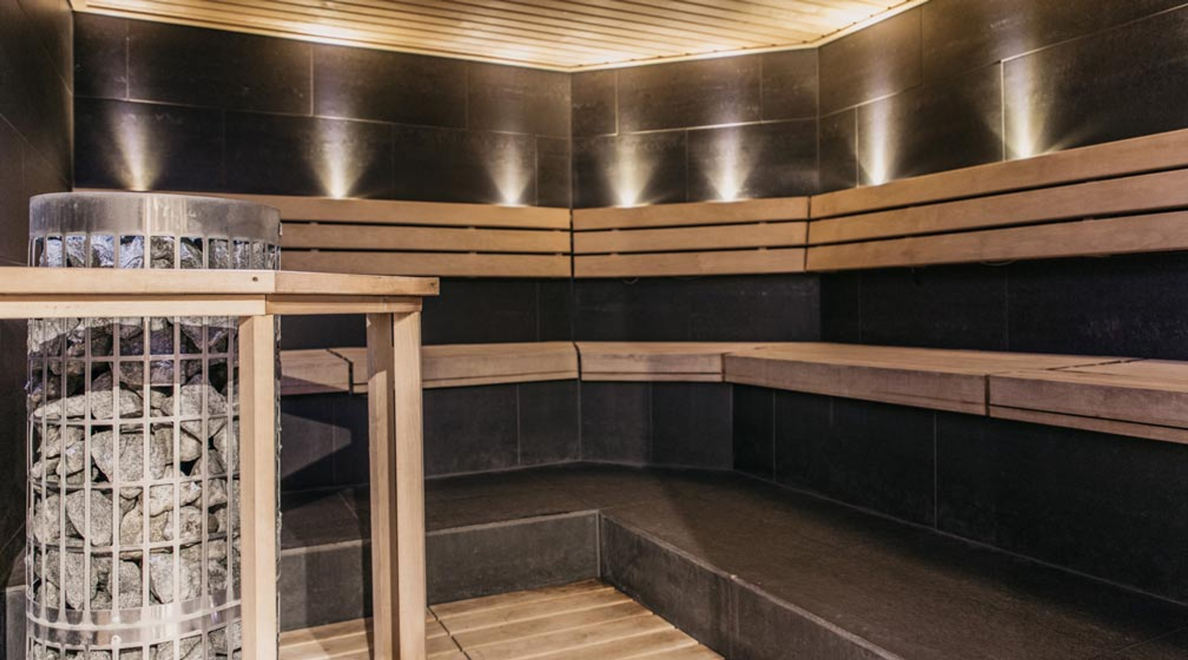 Moderni sauna tummilla laatoitetuilla seinillä, puiset lauteet ja kiuaskivikori nurkassa. Katon puupaneelissa valaistus.