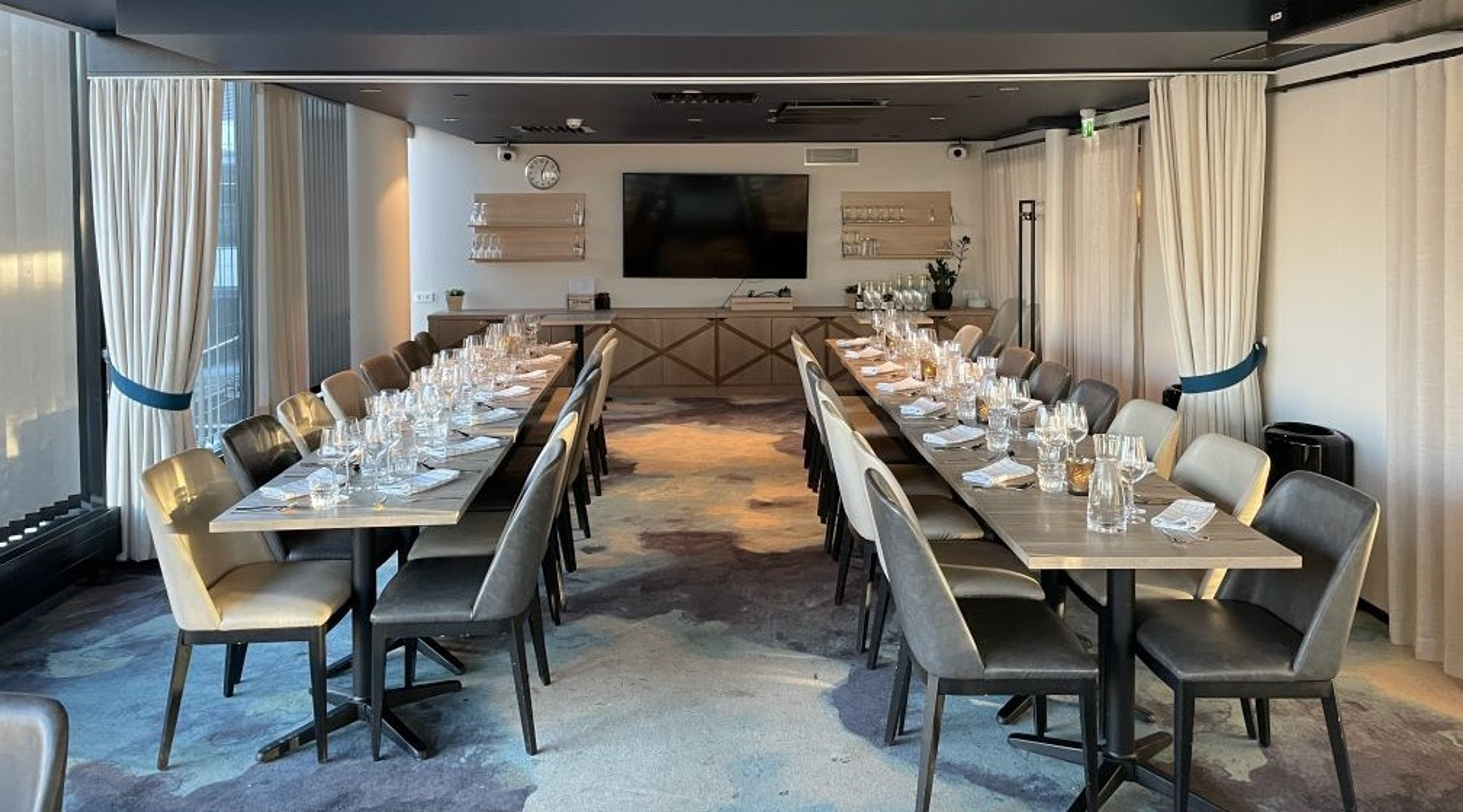 Moderni neuvotteluhuone kahdella pitkällä pöydällä, joihin on katettu lasit ja astiat. Taustalla on televisio ja tarjoilukaluste vaaleilla verhoilla.
