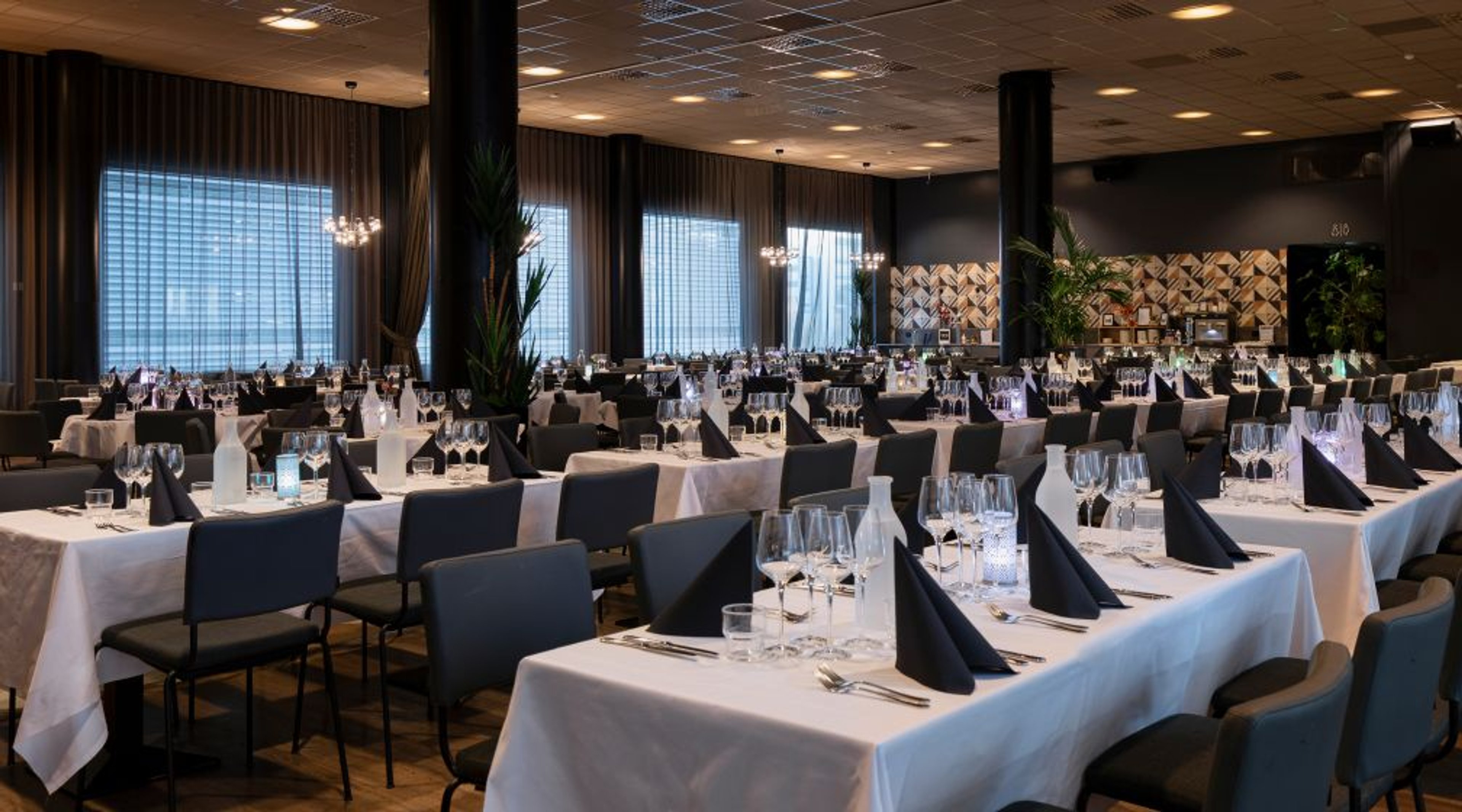 Tilava ravintolasali jossa valkoisilla pöytäliinoilla katettuja pöytiä, sinisiä servettejä ja tummia tuoleja. Taustalla suuret ikkunat ja viherkasveja.