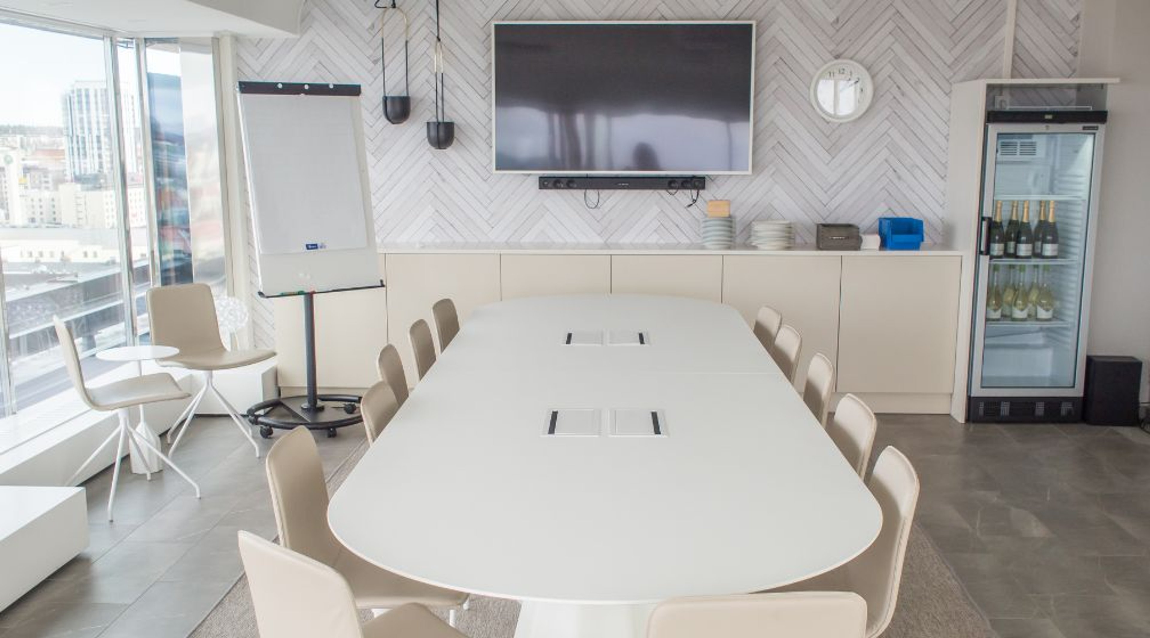 Valoisa neuvotteluhuone, jossa valkoinen pöytä, vaaleat tuolit, fläppitaulu, televisio ja juomakaappi. Isot ikkunat tarjoavat kaupunkinäkymän.