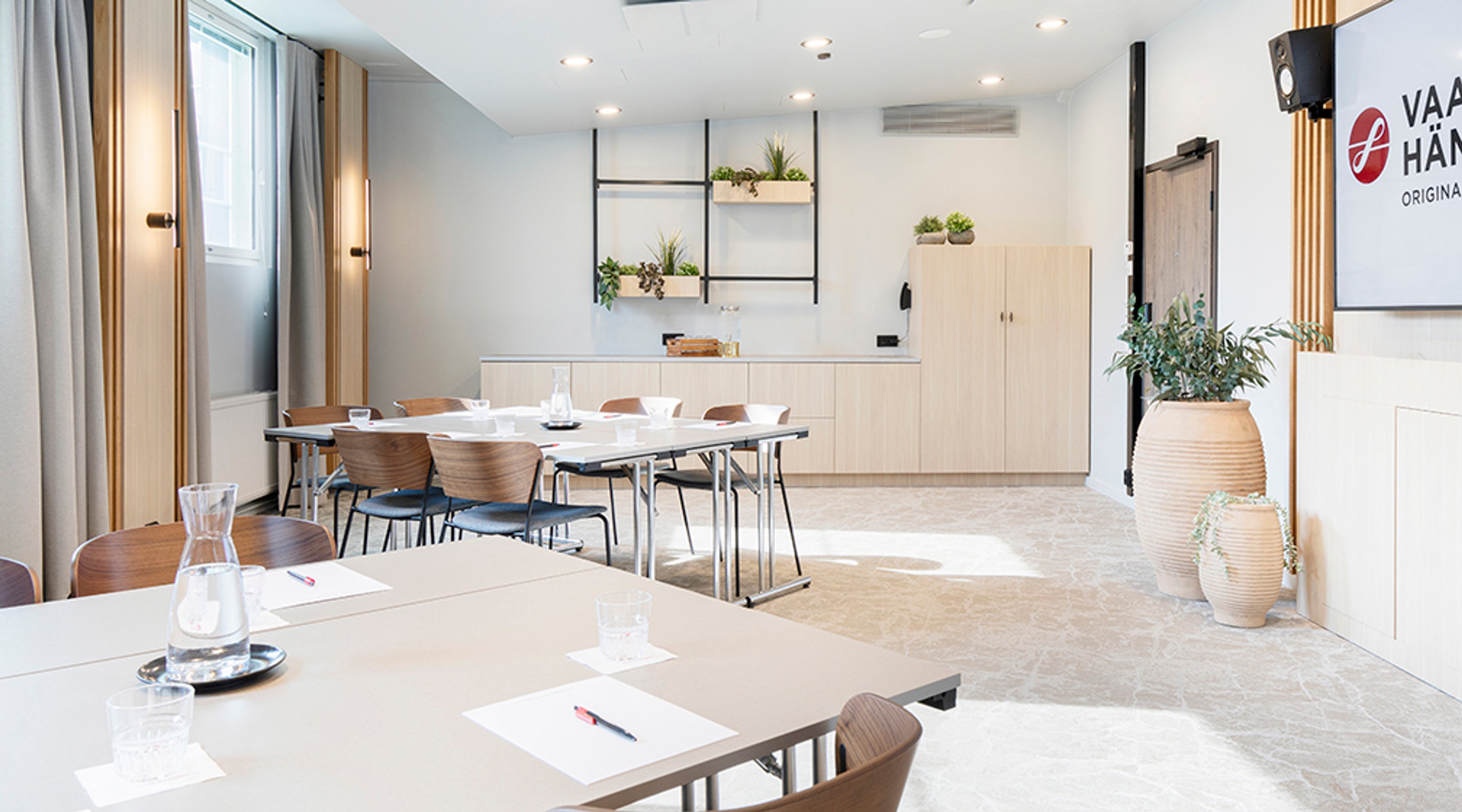 Valoisa kokoustila, jossa modernit puutuolit ja pöydät, valkoinen keittiökulmaus, seinähyllyt kasveille ja tasainen marmorimainen lattia.