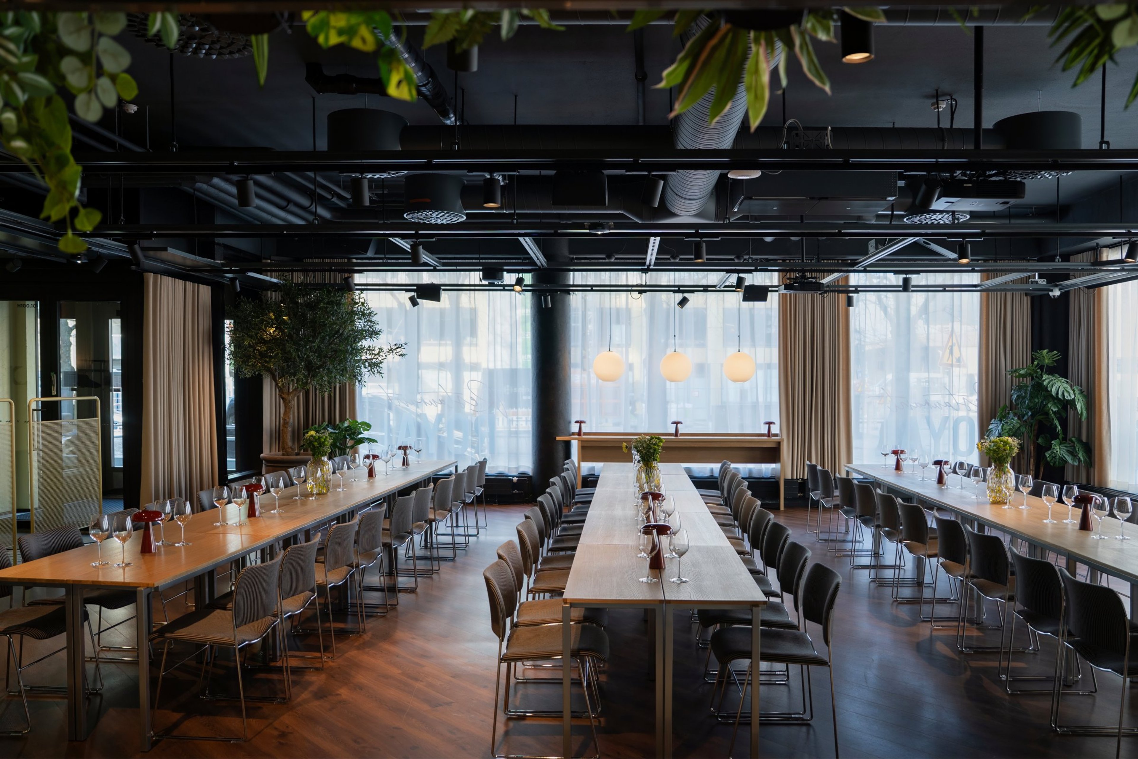 Moderni ravintolasali kolmella pitkällä pöydällä, puulattia, isot ikkunat verhoineen ja viherkasveilla. Tasainen lattia, avara tila.