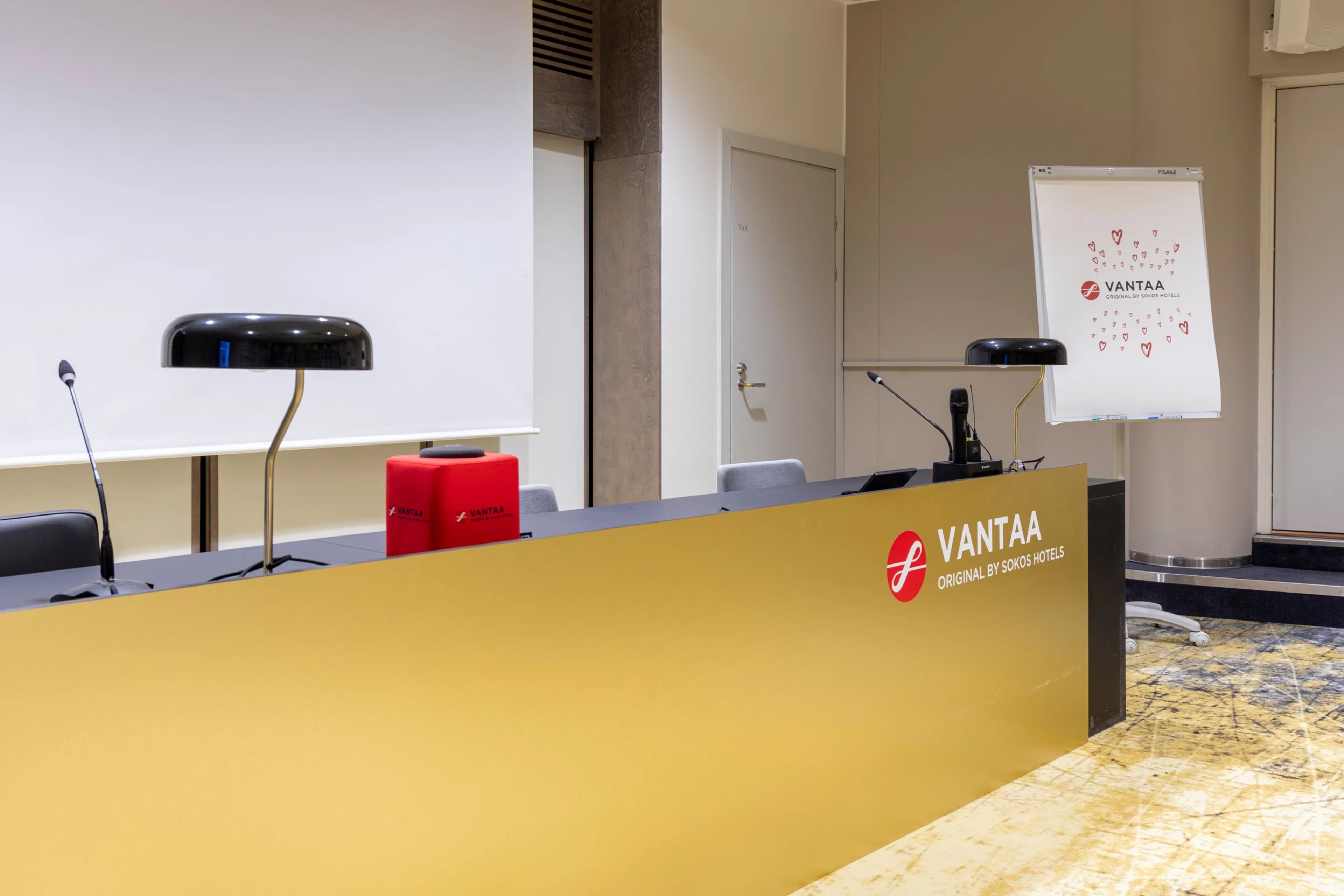 Yellow reception desk at Vantaa hotel. Black desk lamp and red box on the counter. White door and a flip chart visible in the background.