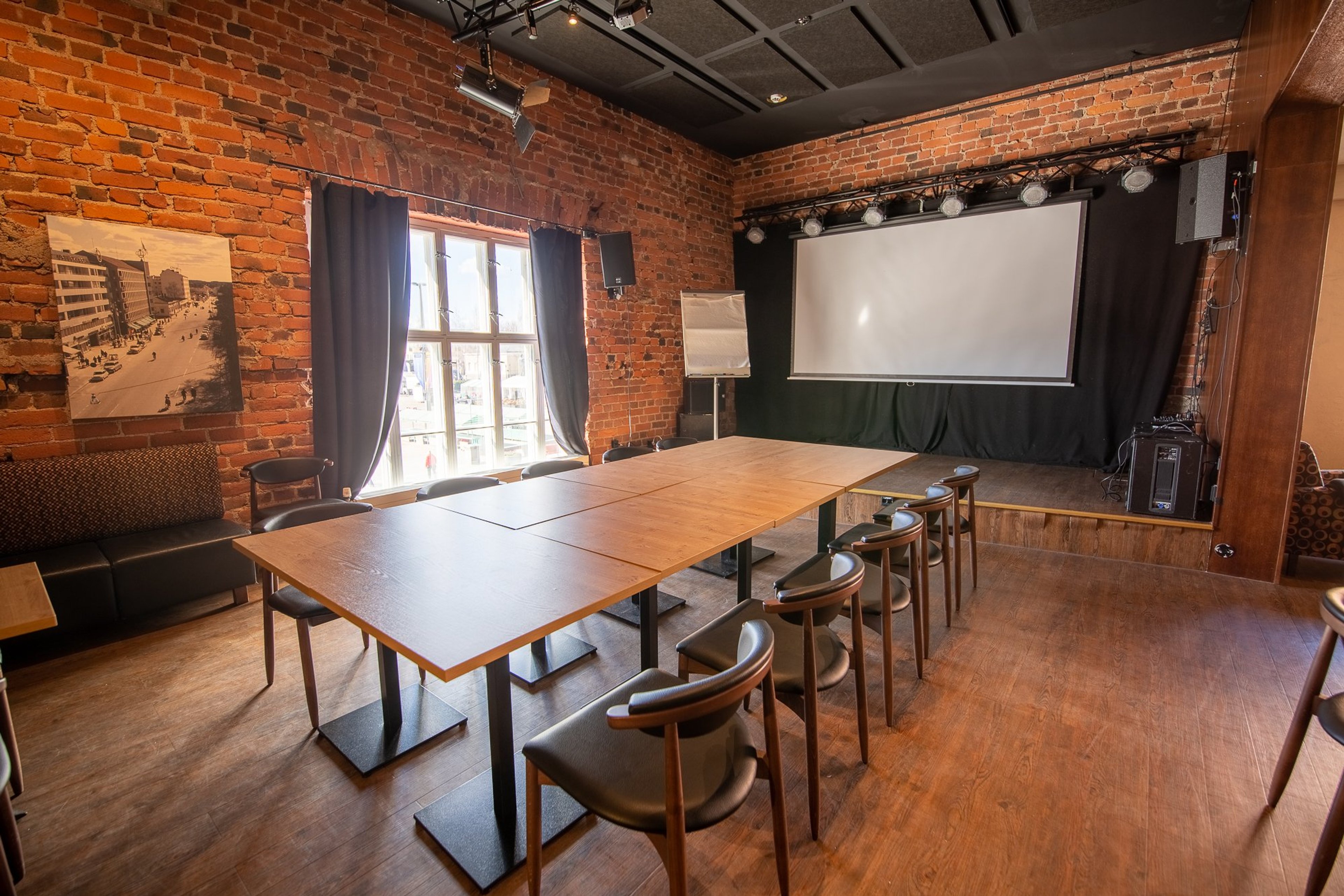 Kokoushuone tiiliseinillä, jossa suuri puupöytä tuoleineen, projektori, valkokangas ja ikkunat verhoilla. Lattia on kovaa puumateriaalia.