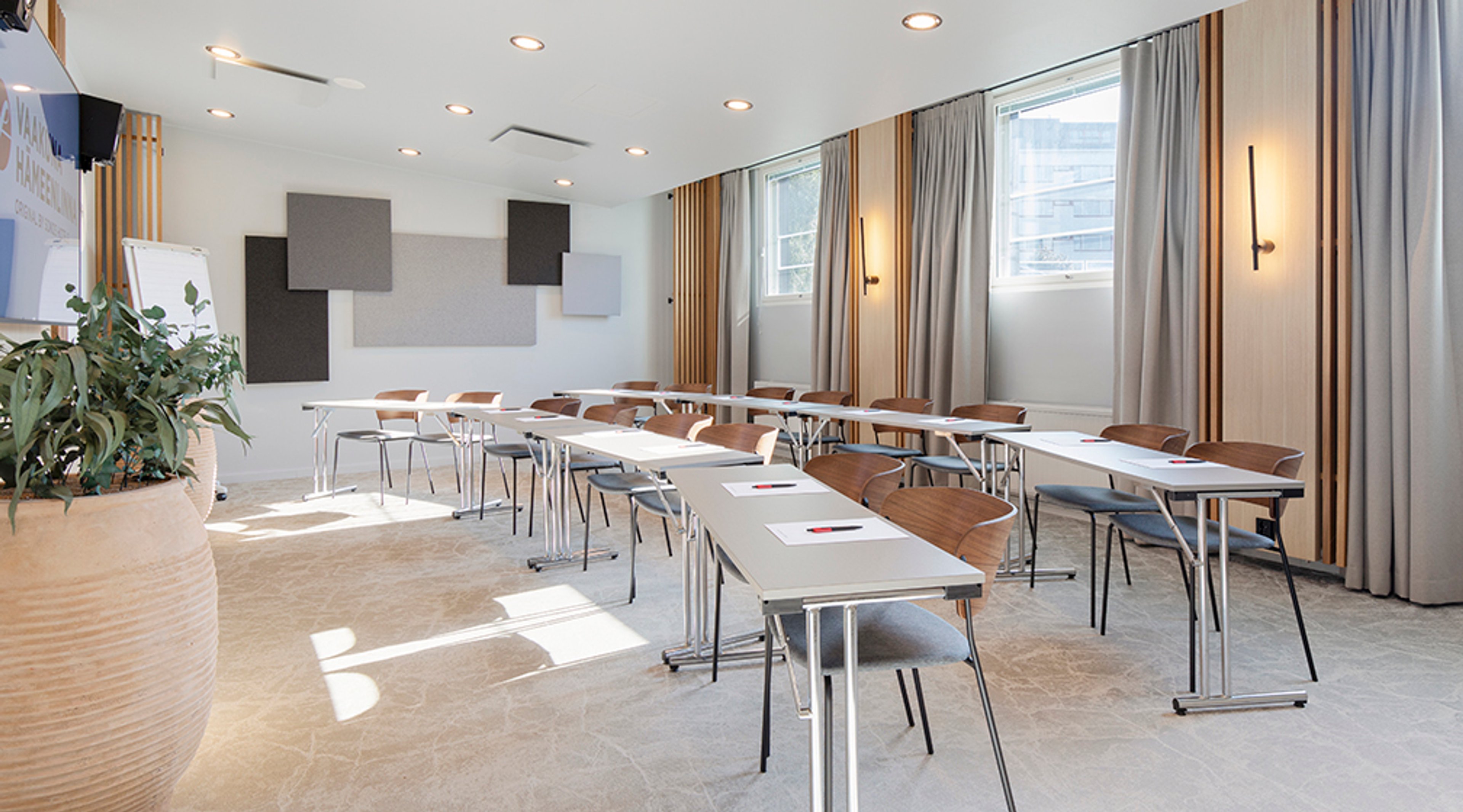 Valoisa kokoustila, jossa pöydät asetettu luokkamuodostelmaan. Huoneessa on harmaat ja beigen sävyiset akustiikkapaneelit seinällä ja kasvi nurkassa.