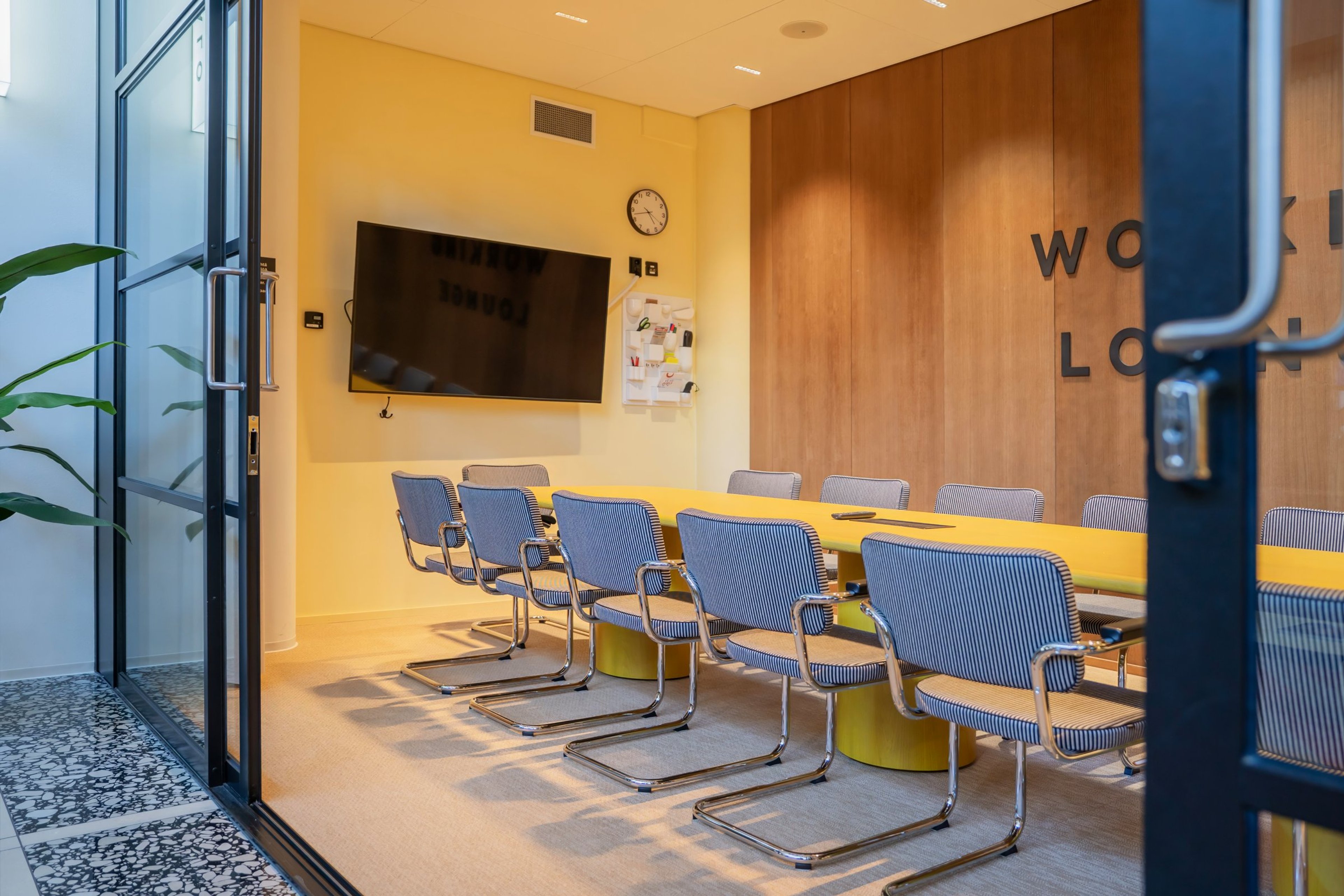 Moderni neuvotteluhuone keltaisella pöydällä, siniraidallisilla tuoleilla ja seinälle asennetulla näytöllä. Leveät ovet ja tasainen lattia.