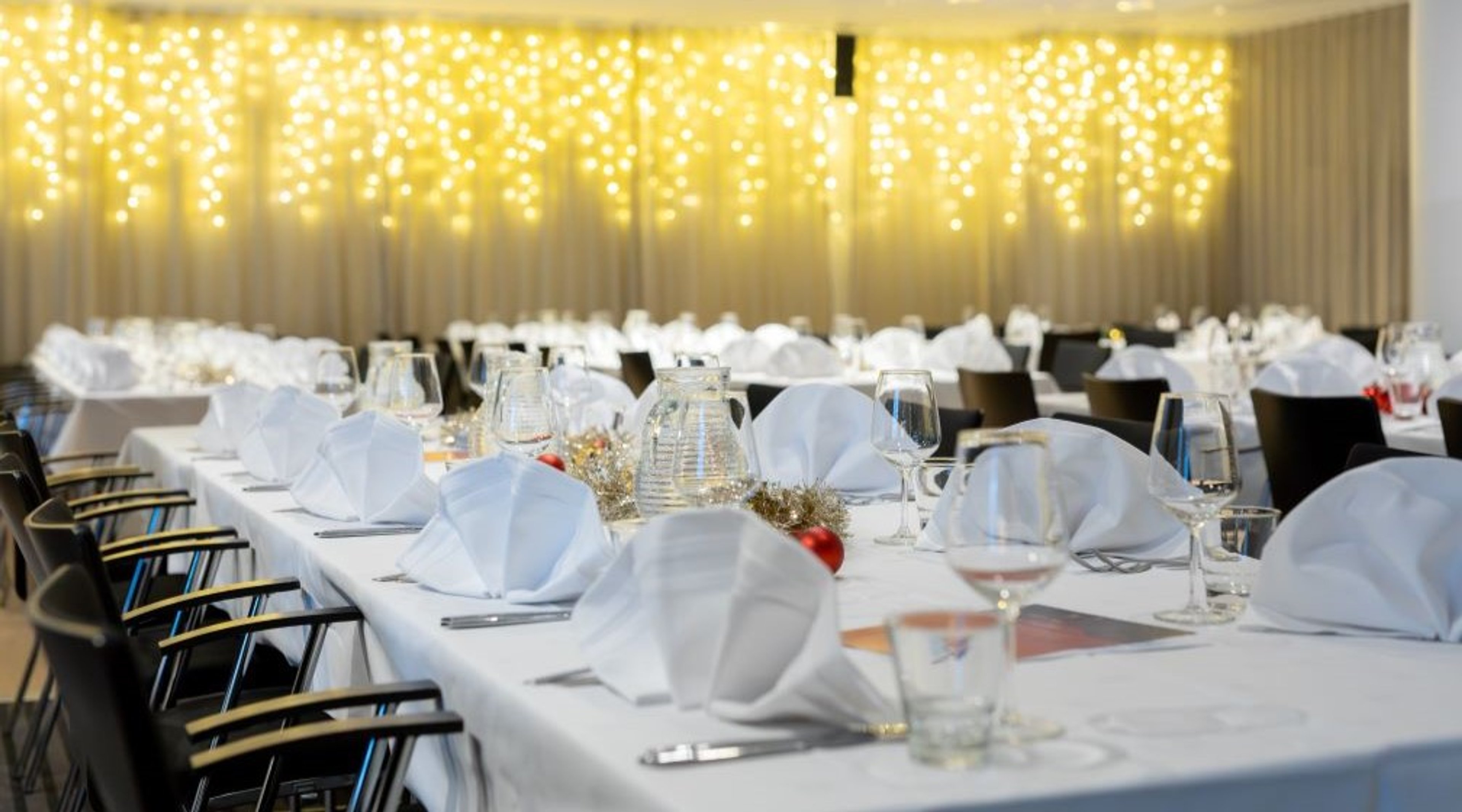 Juhlallisesti katettu ruokailutila, jossa valkoiset pöytäliinat ja taiteltuina lautasliinat. Taustalla seinäverhot, joita valaisevat koristevalot.