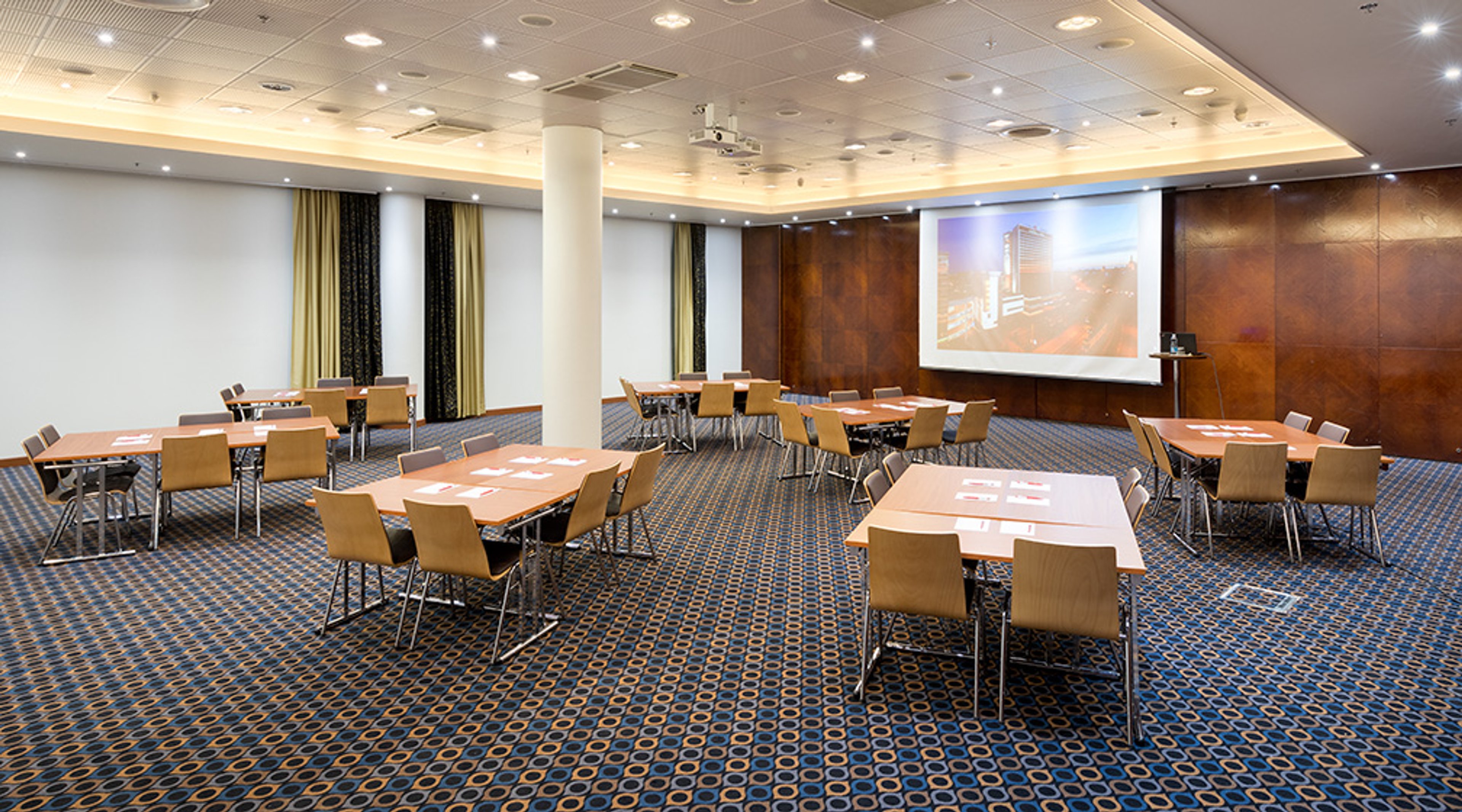 Konferenssihuone, jossa on pöytiä neliön muotoisessa järjestyksessä, projektorikangas seinällä ja kuvioitu kokolattiamatto.