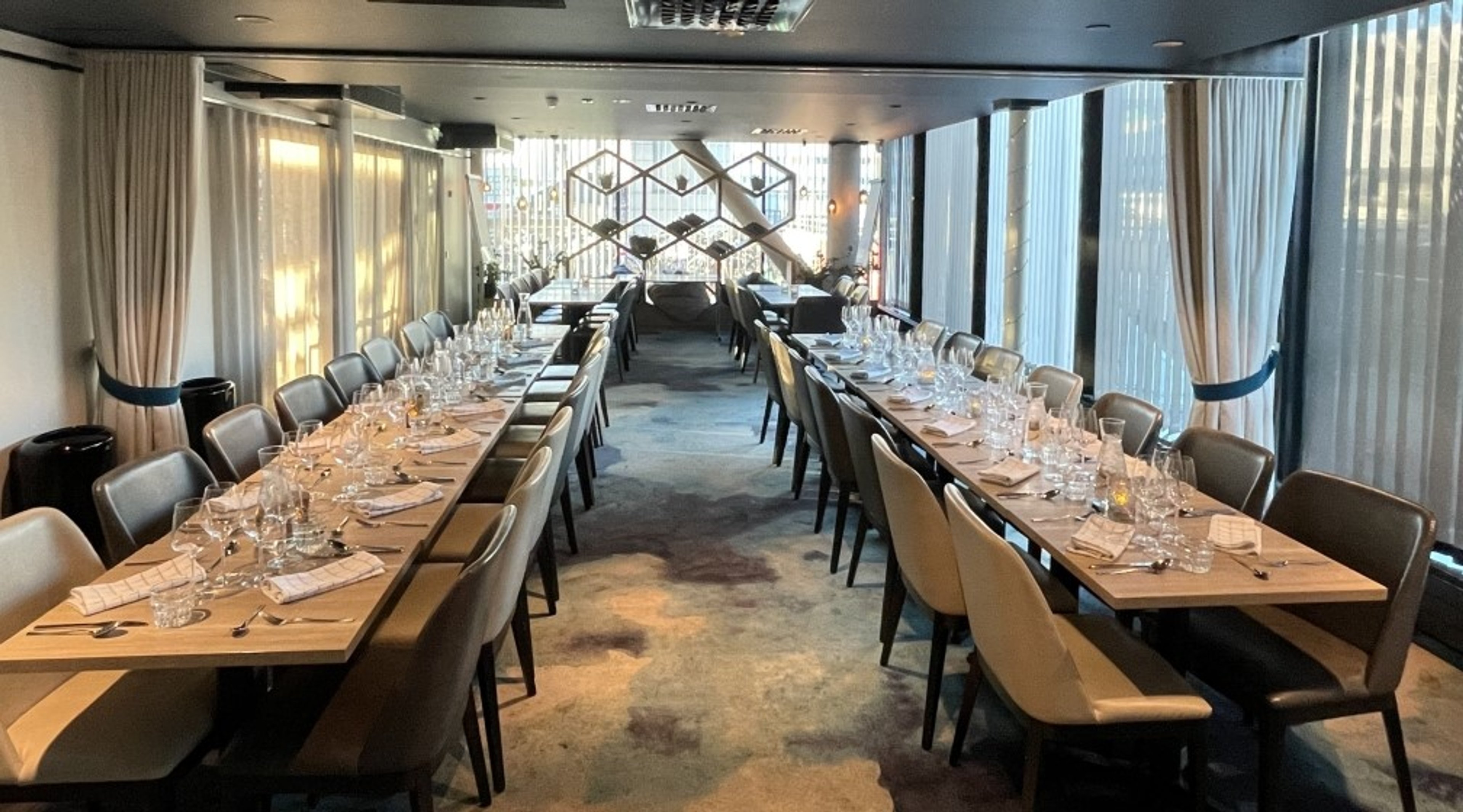 Ravintolan yksityinen tila, jossa kaksi pitkää pöytää kattauksella. Huone on valoisa, ikkunoilla pehmeiden verhojen takana ja koristeellisella tilanjakajalla.