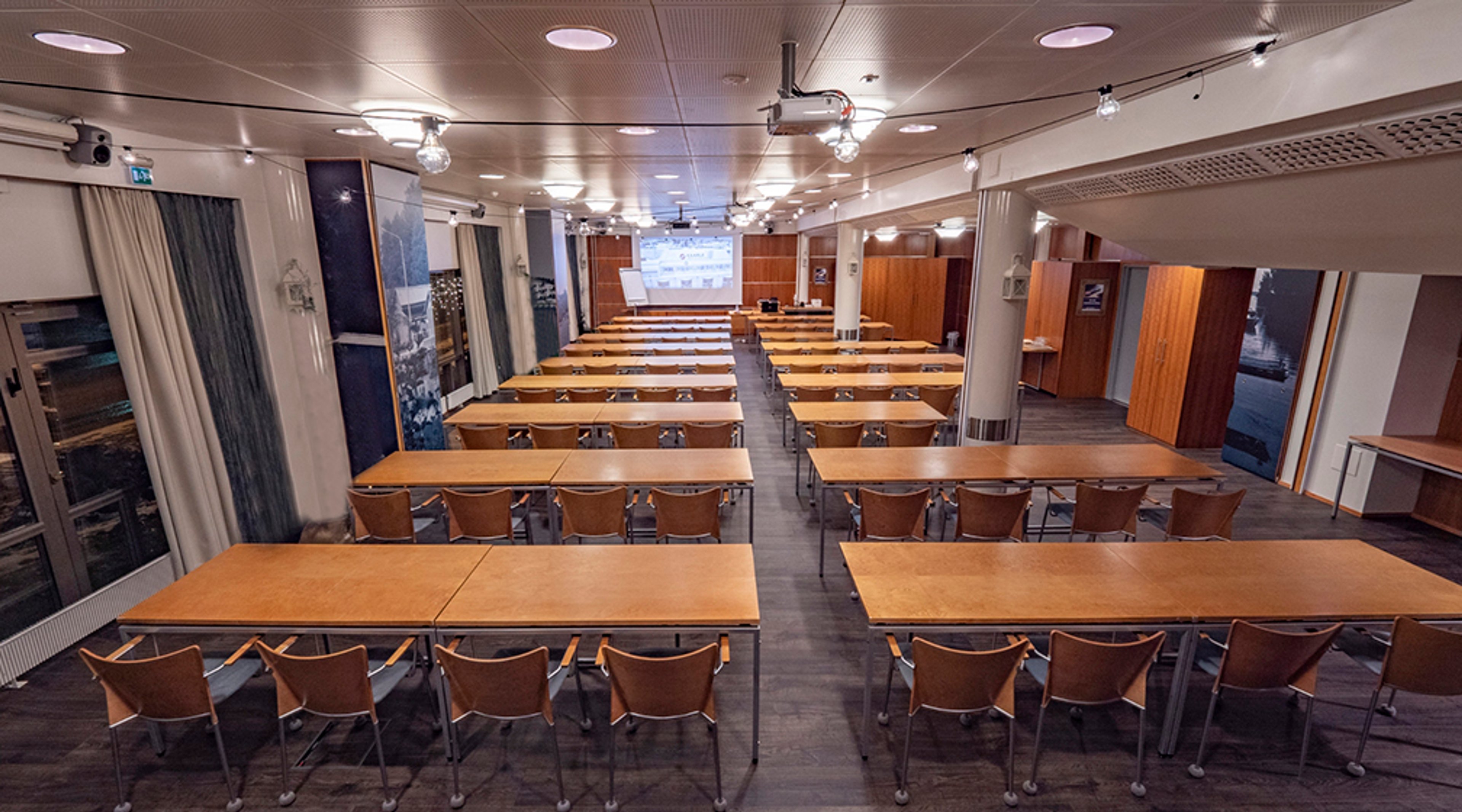 Conference room with rows of wooden tables and chairs. Projection screen at the front and windows with curtains along the walls. Floor is dark and hard.