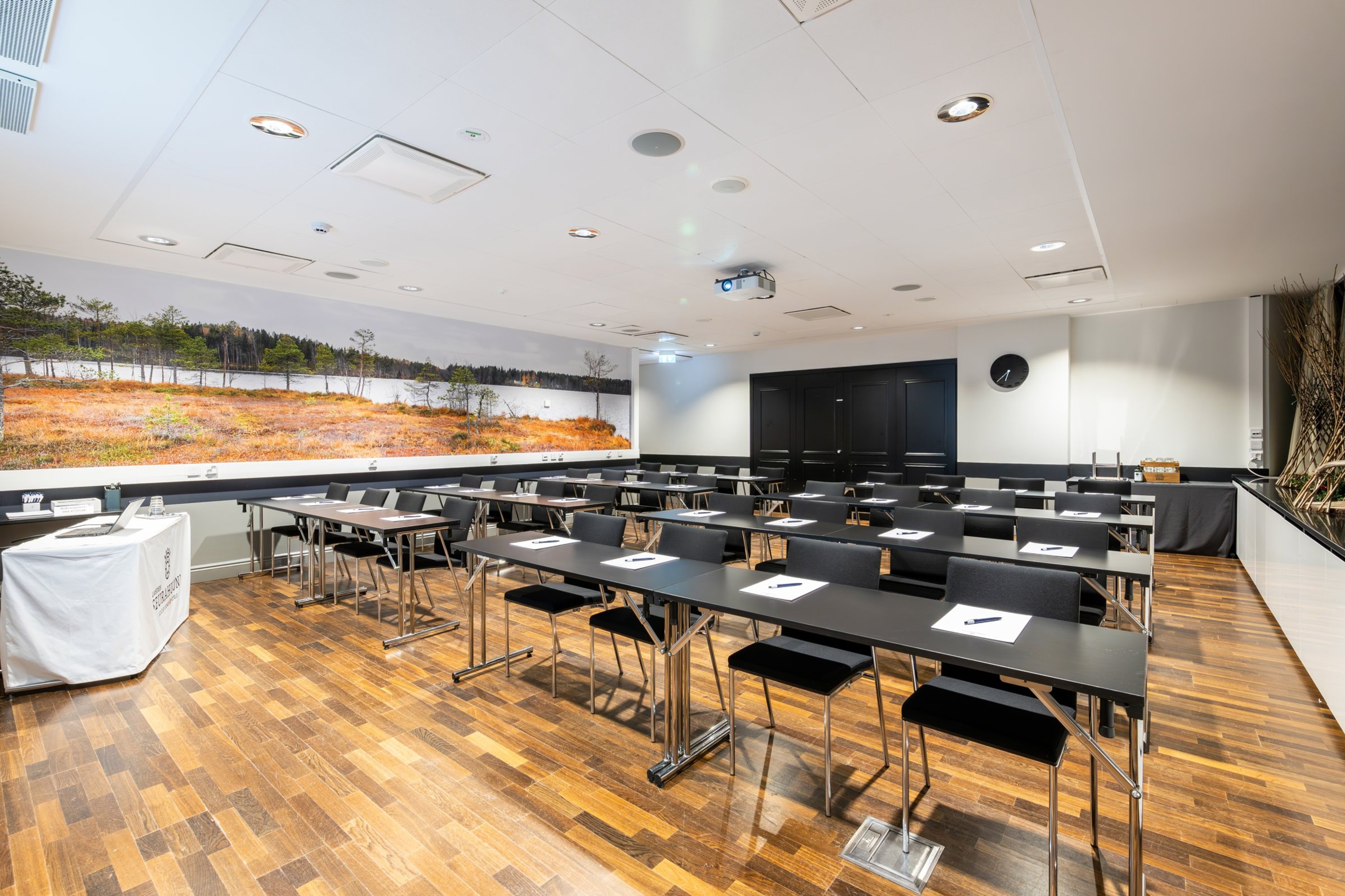 Moderni kokoushuone puulattialla, mustilla pöydillä ja tuoleilla. Seinällä suuri maisemakuva suoalueesta. Tila on hyvin valaistu.