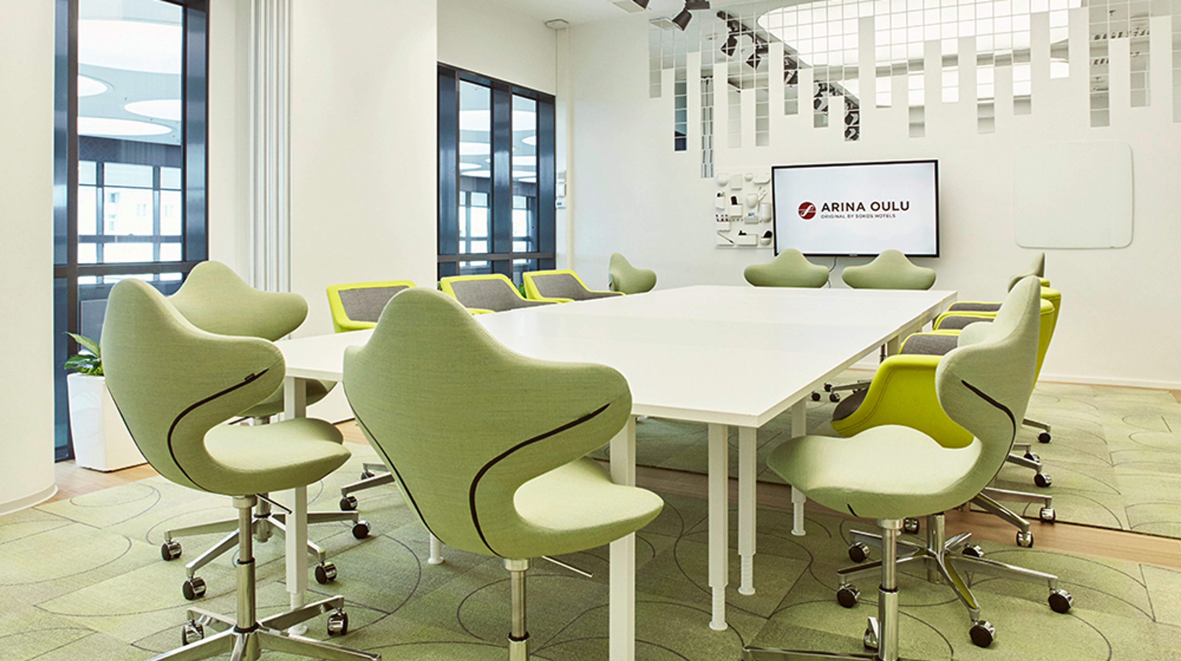 Moderni neuvottelutila valkoisella pöydällä ja tyylikkäillä vaaleanvihreillä tuoleilla. Taustalla näyttö ja lasiseinät.