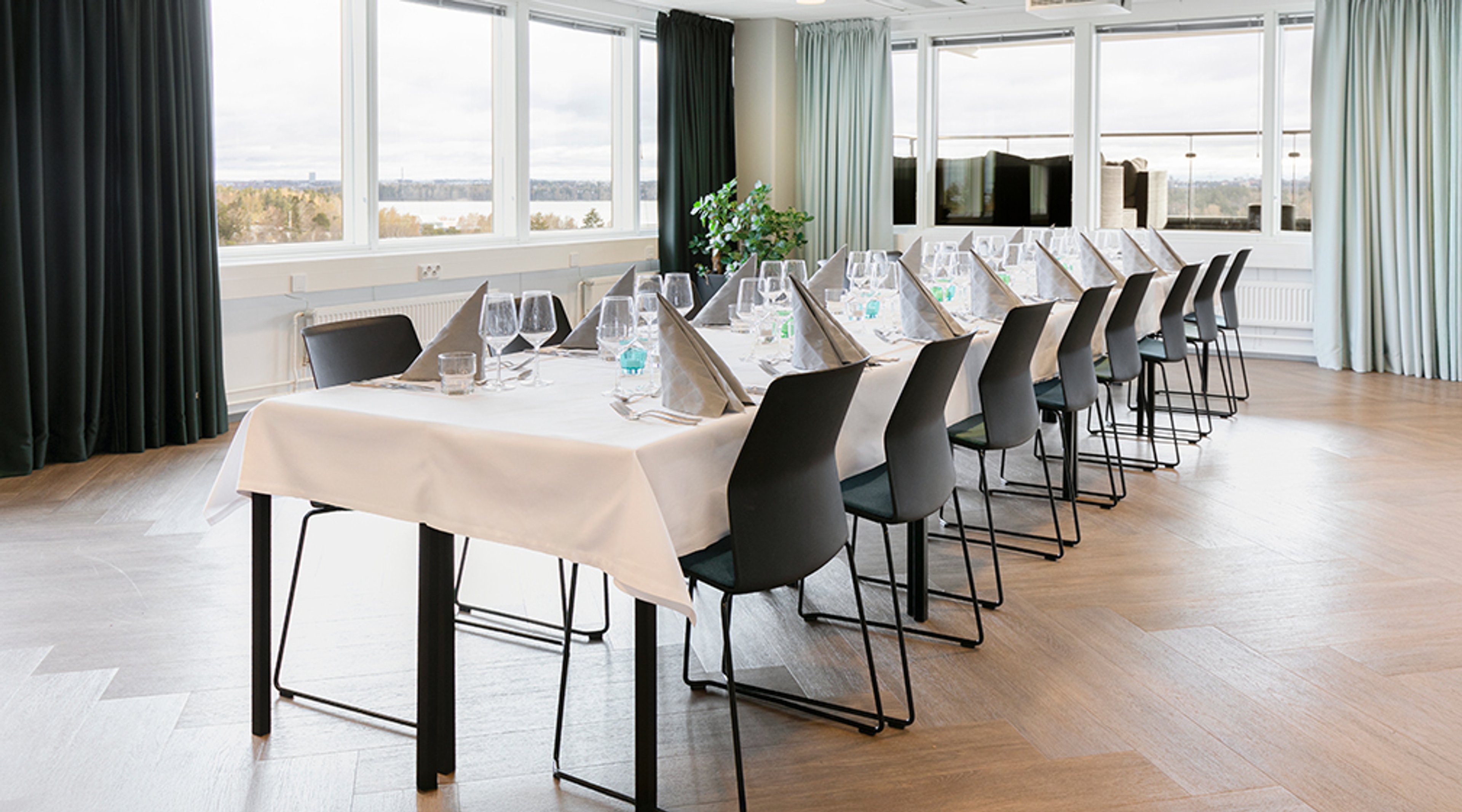 Spacious meeting room with bright windows. Long table with white tablecloth set with wine glasses. Soft wooden floor and black chairs.