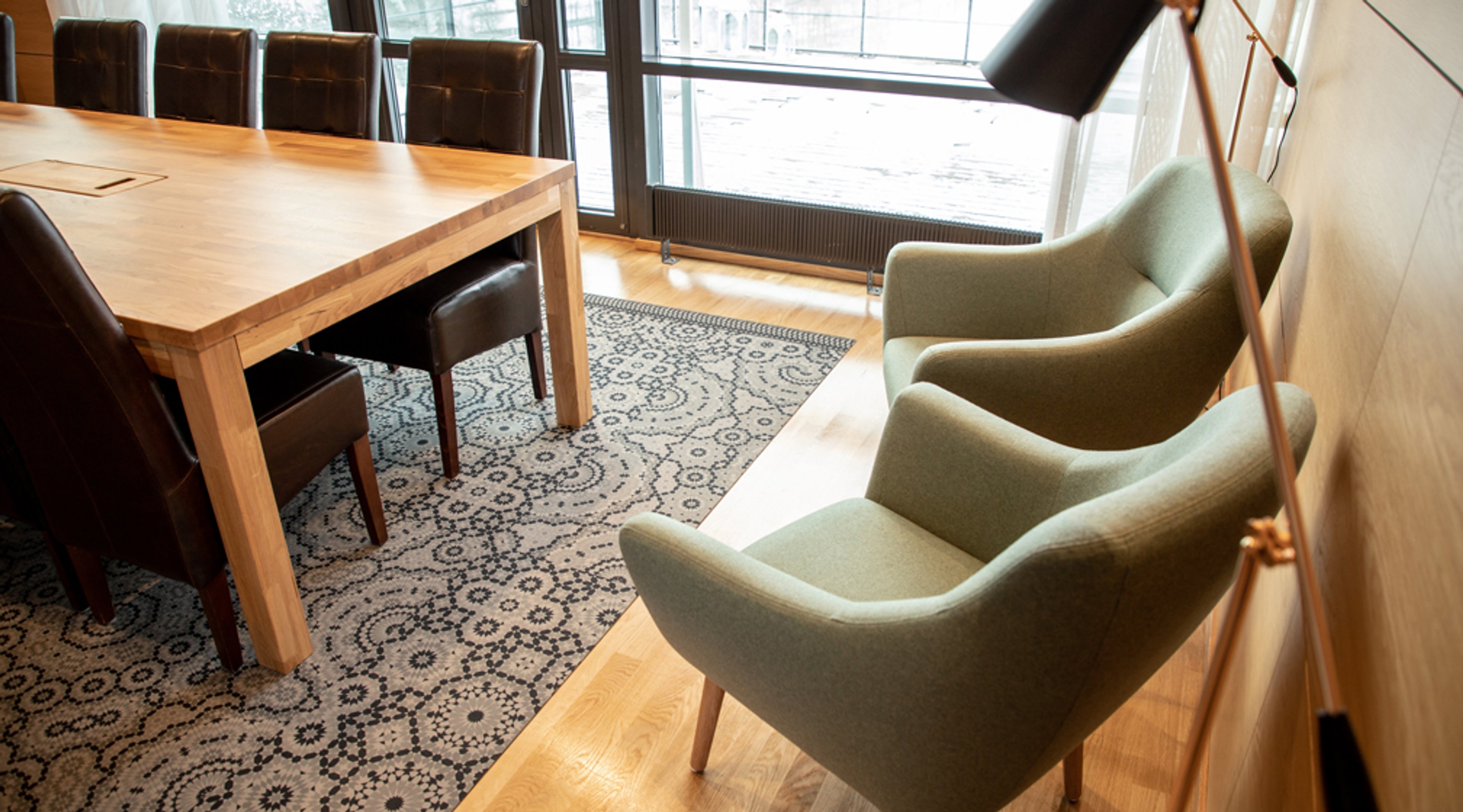 Kokoushuone, jossa puinen pöytä, tummanruskeita tuoleja, vaaleanvihreitä nojatuoleja ja kuviollinen matto vaalealla puulattialla.