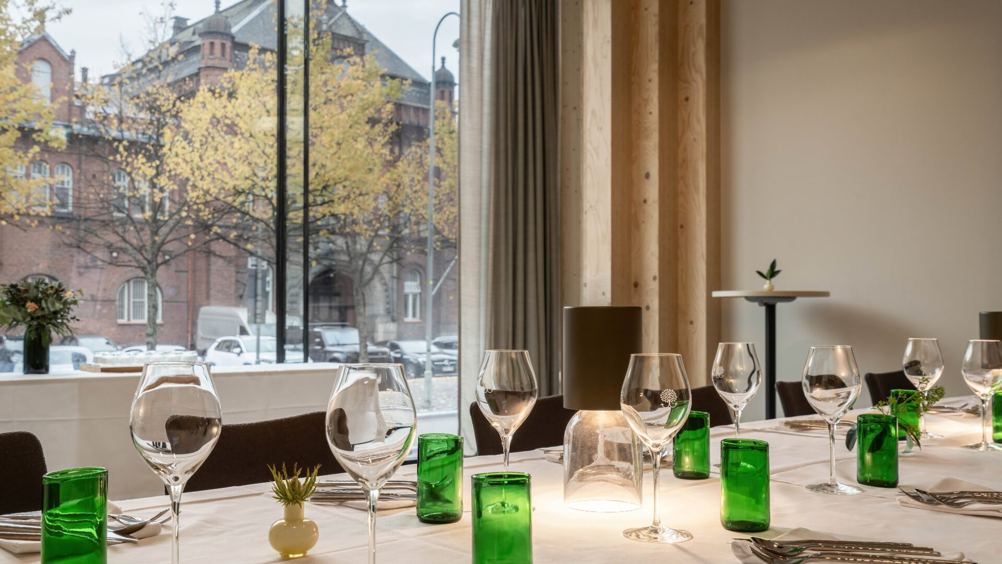 Pöytä viinilaseineen ja vihreine juomalaseineen. Isosta ikkunasta avautuu näkymä historialliseen tiilirakennukseen ja keltalehtisiin puihin.
