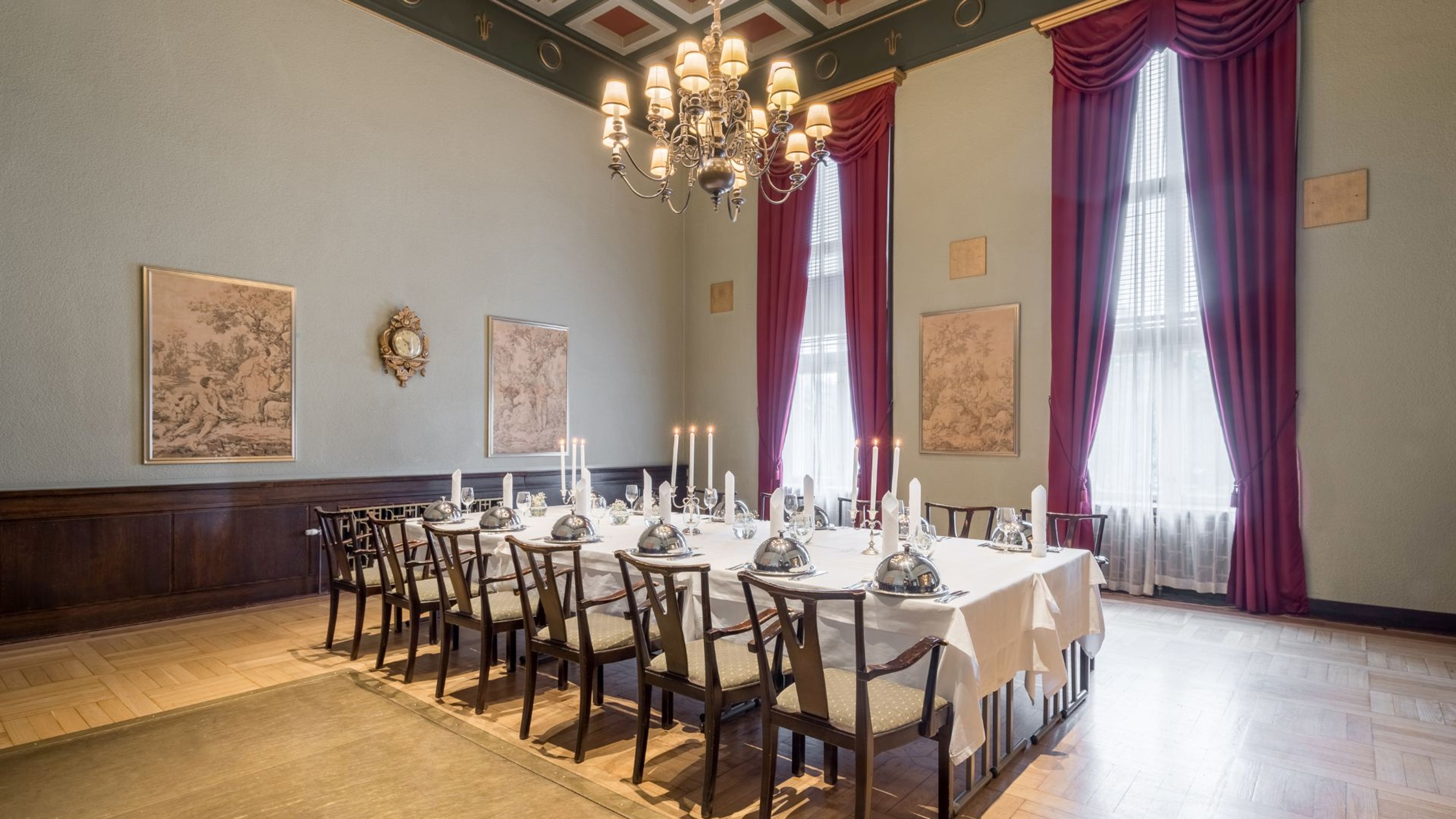 Elegantti juhlahuone, jossa pitkä pöytä kynttilöillä ja hopeakuvuilla, punaiset verhot, koristeellinen kattokruunu ja puulattia.
