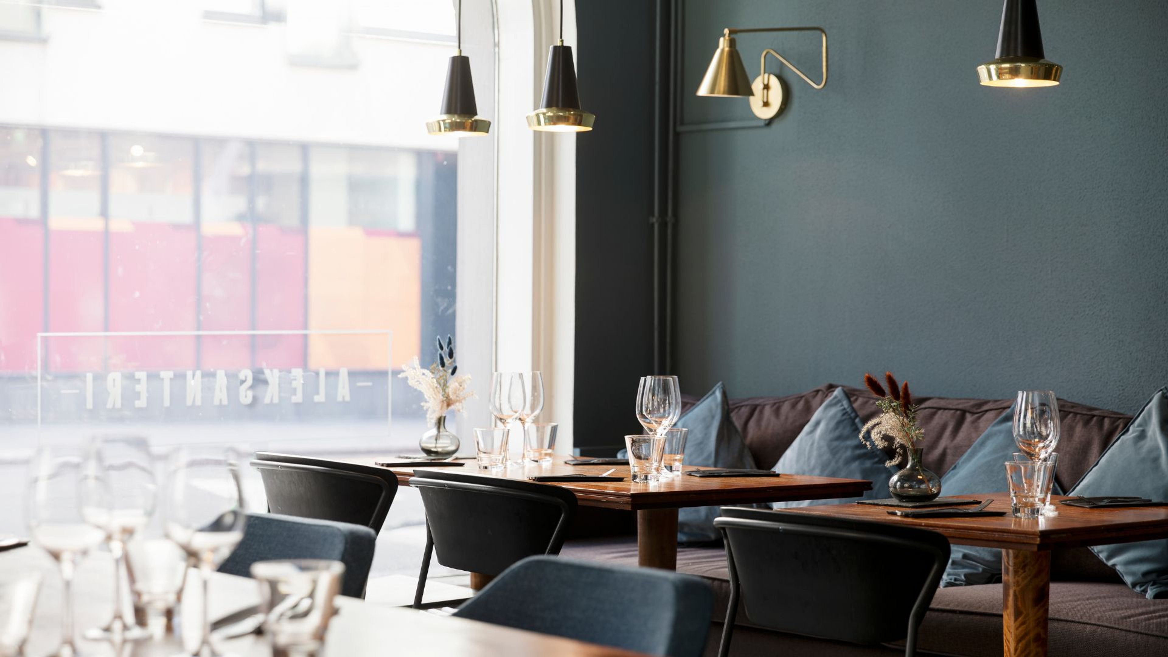 Tyylikkään ravintolan sisustus tummanvihreillä seinillä, puupöydillä ja modernilla valaistuksella. Lasiset ikkunat tuovat luonnonvaloa tilaan.