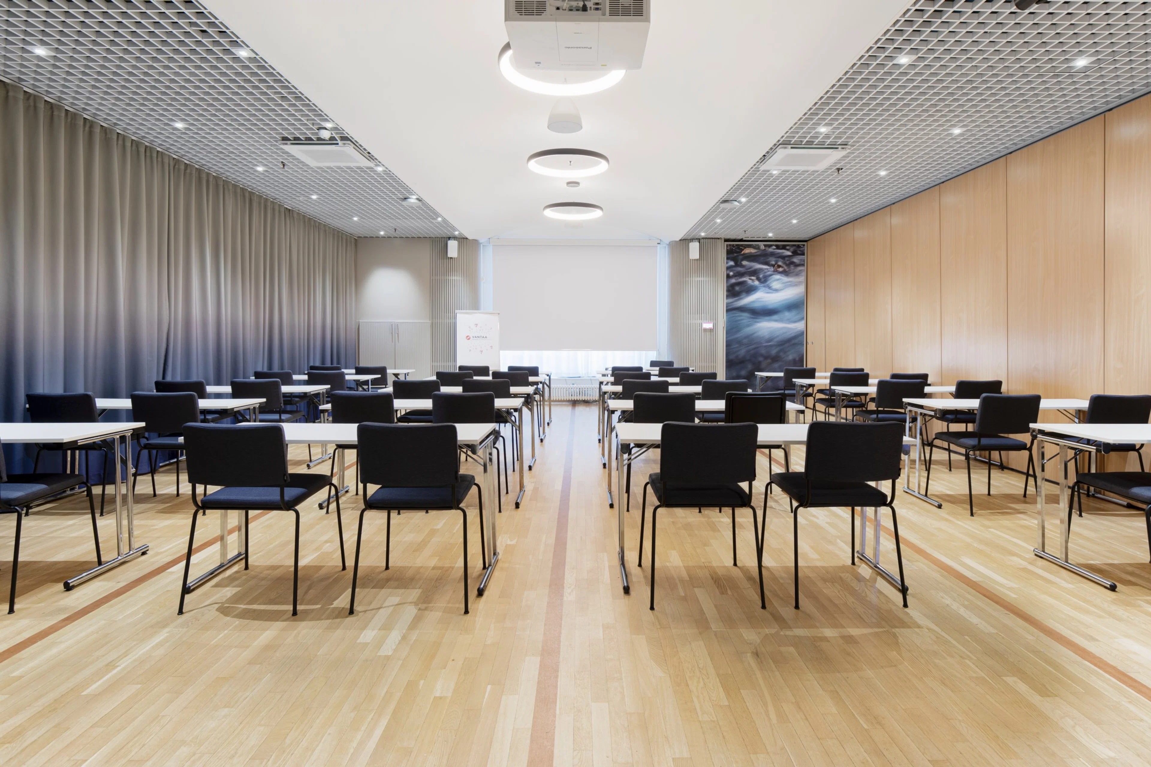 Valoisa kokoustila jossa riveihin järjestettyjä pöytiä ja mustia tuoleja. Puulattia, harmaat verhot ja vaaleat seinät. Tasainen valaistus katossa.