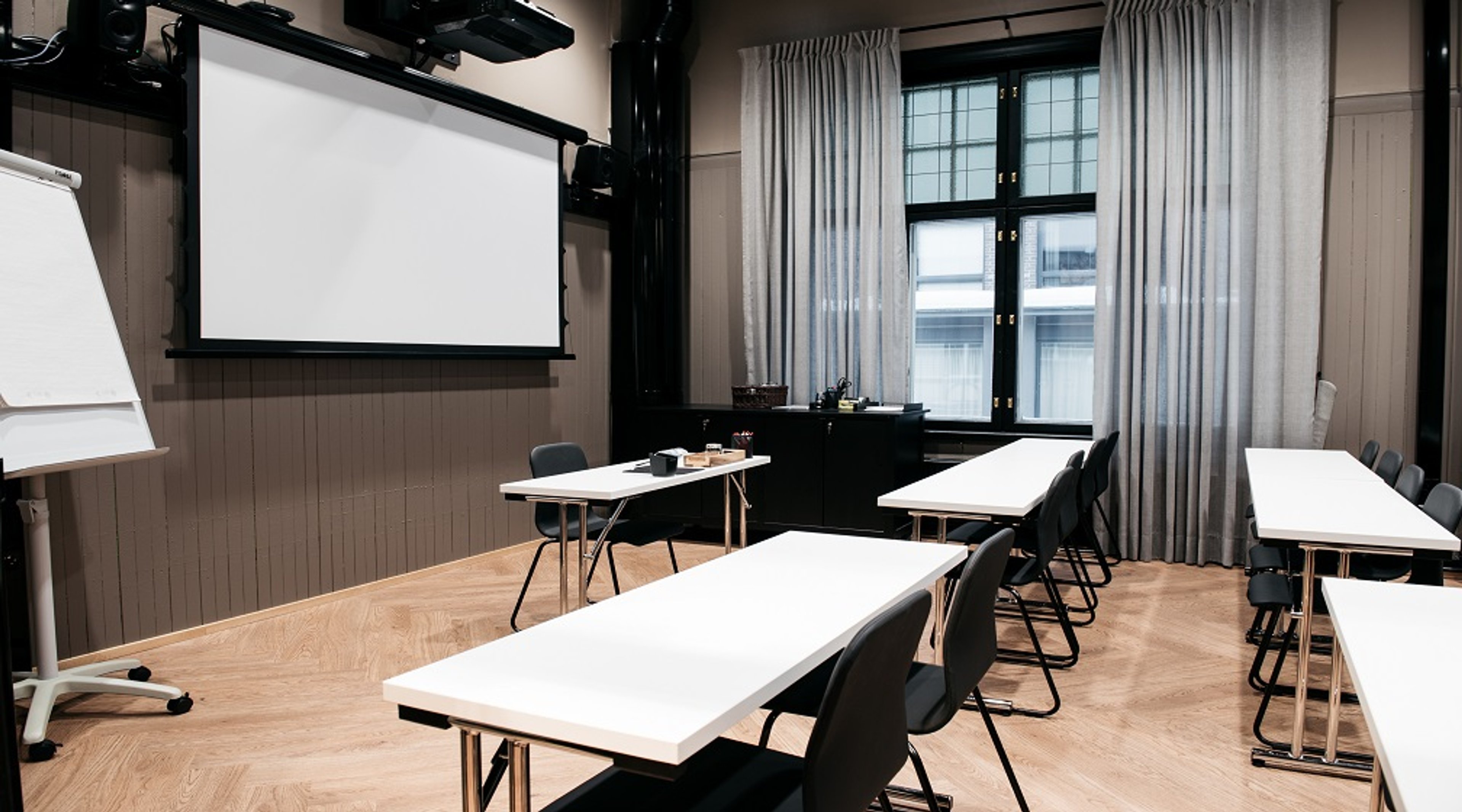 Kokoushuone, jossa on valkoinen projektoriruutu, useita valkopintaisia pöytiä ja tuoleja sekä vaalea puulattia ja fläppitaulu nurkassa.