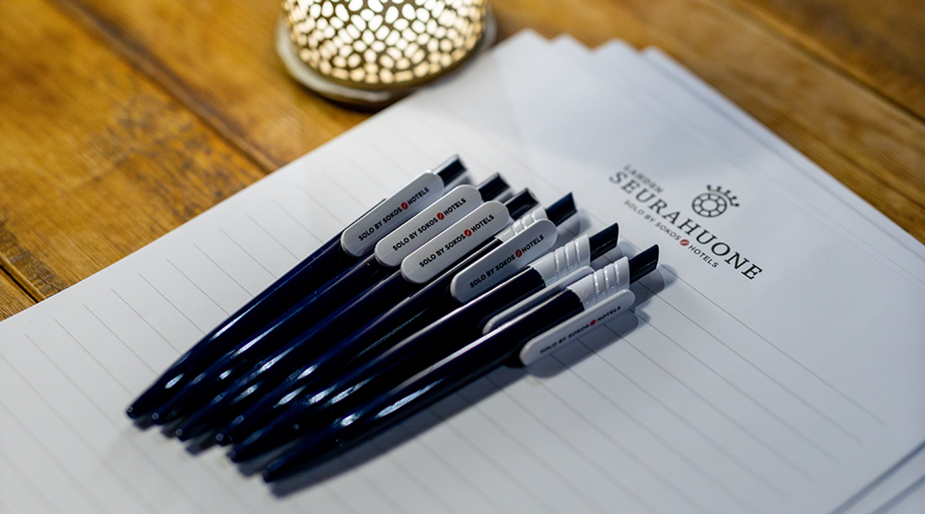 Kuusi sinistä hotellikynää aseteltuna riviin valkoiselle paperille puisella pöydällä, koristeellinen kynttilä taustalla.