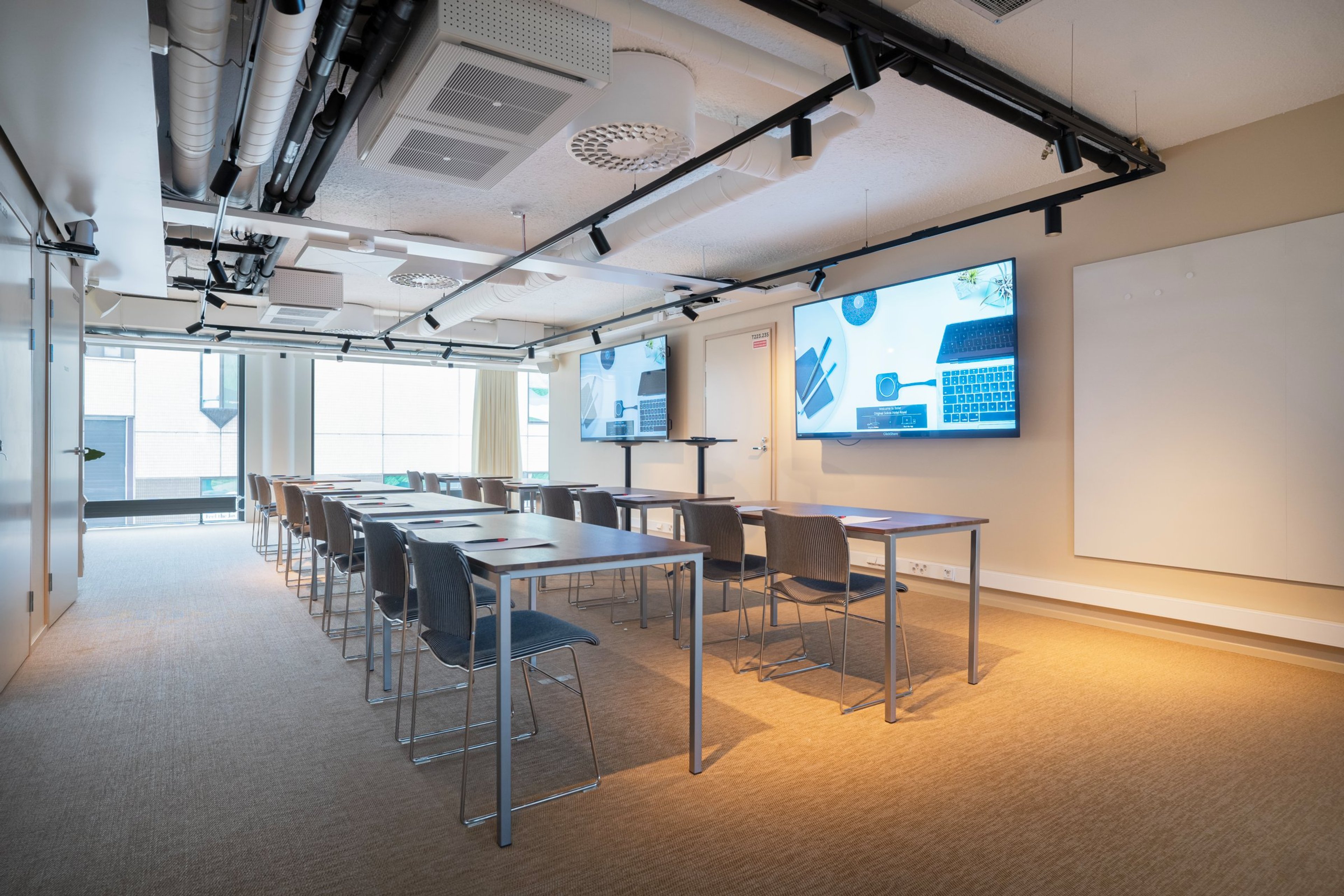Moderni kokoustila jossa pöydät ja tuolit rivissä, kaksi suurta näyttöä seinällä ja kattoon upotetut valot. Lattia on tasainen ja kulkuväylä leveä.