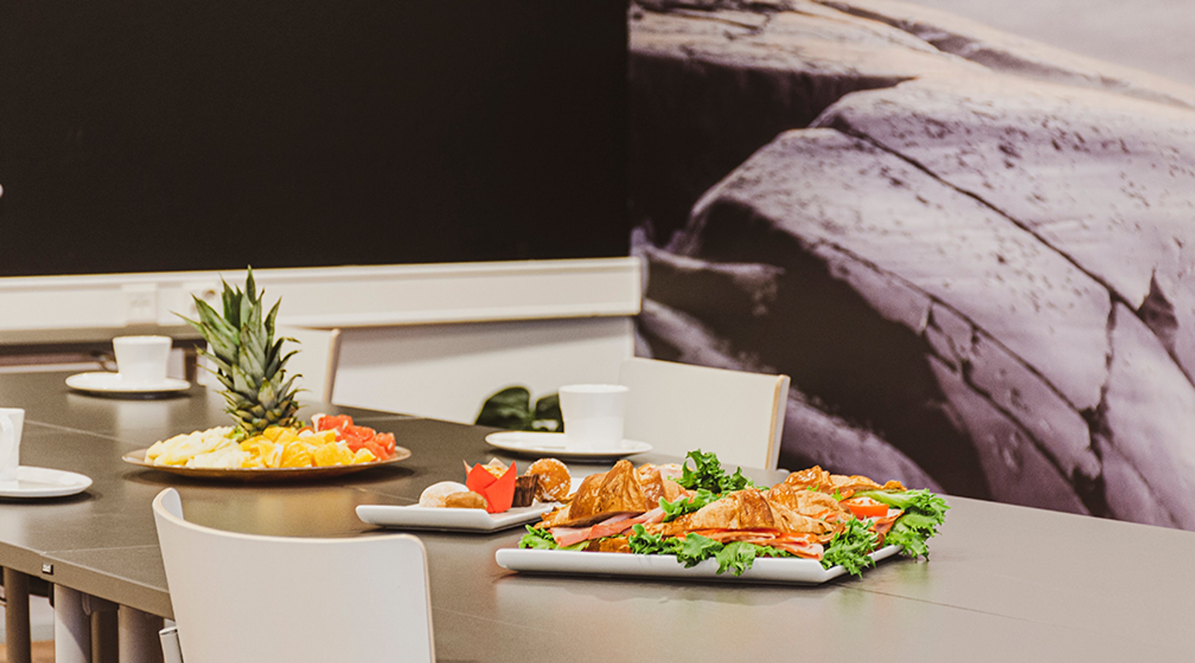 Aamiaispöytä, jossa tarjolla croissantteja, vihersalaattia sekä hedelmälautanen ananaksen kera. Tarjoilu modernissa tilassa.