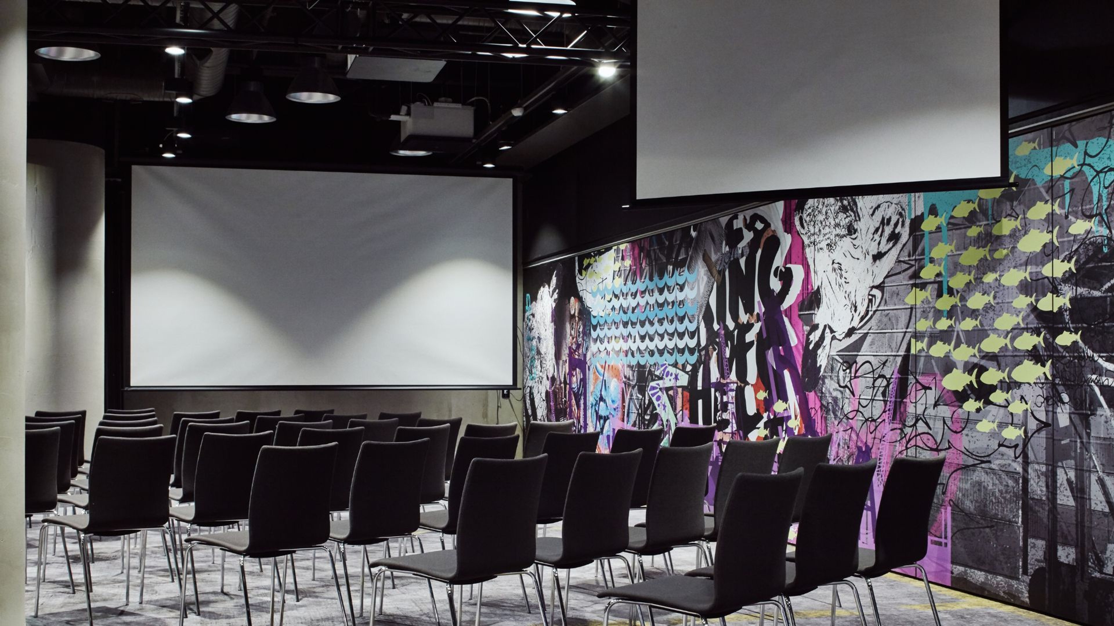 Moderni auditorio mustilla tuoleilla, valkoinen valkokangas edessä ja värikäs graffitityyppinen seinämaalaus oikealla.