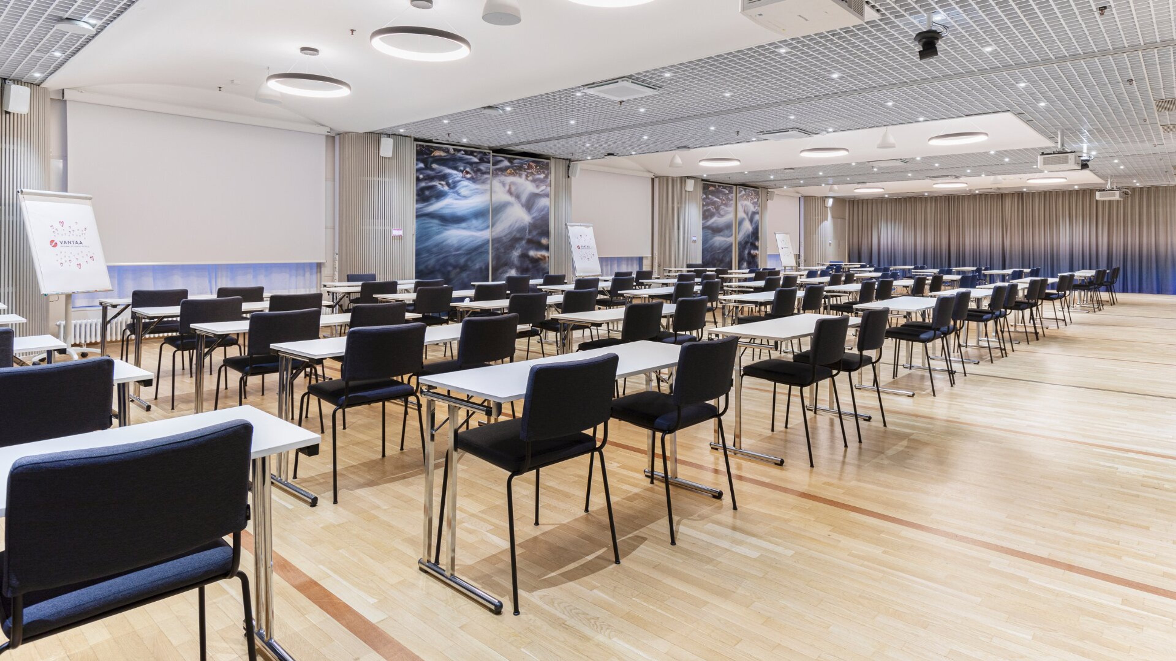 Modern conference room with bright wooden floor, black chairs and white tables arranged in rows. Nature-themed wall art and flipcharts visible along the sides.