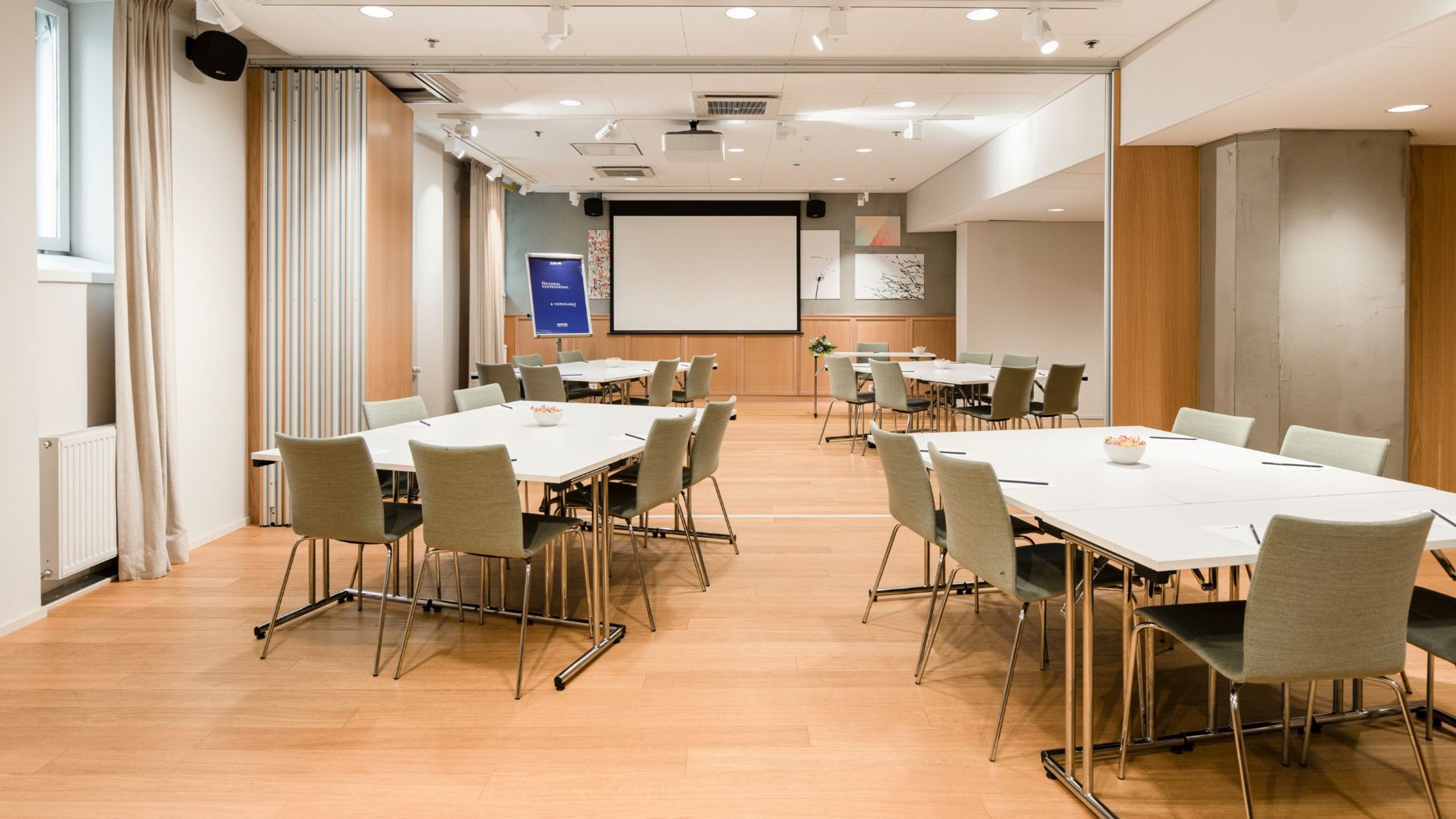 Moderni kokoustila, jossa vaalea puulattia, useita pöytiä, harmaita tuoleja ja valkokangasnäyttö. Tilassa on hyvä valaistus ja tasainen lattia.