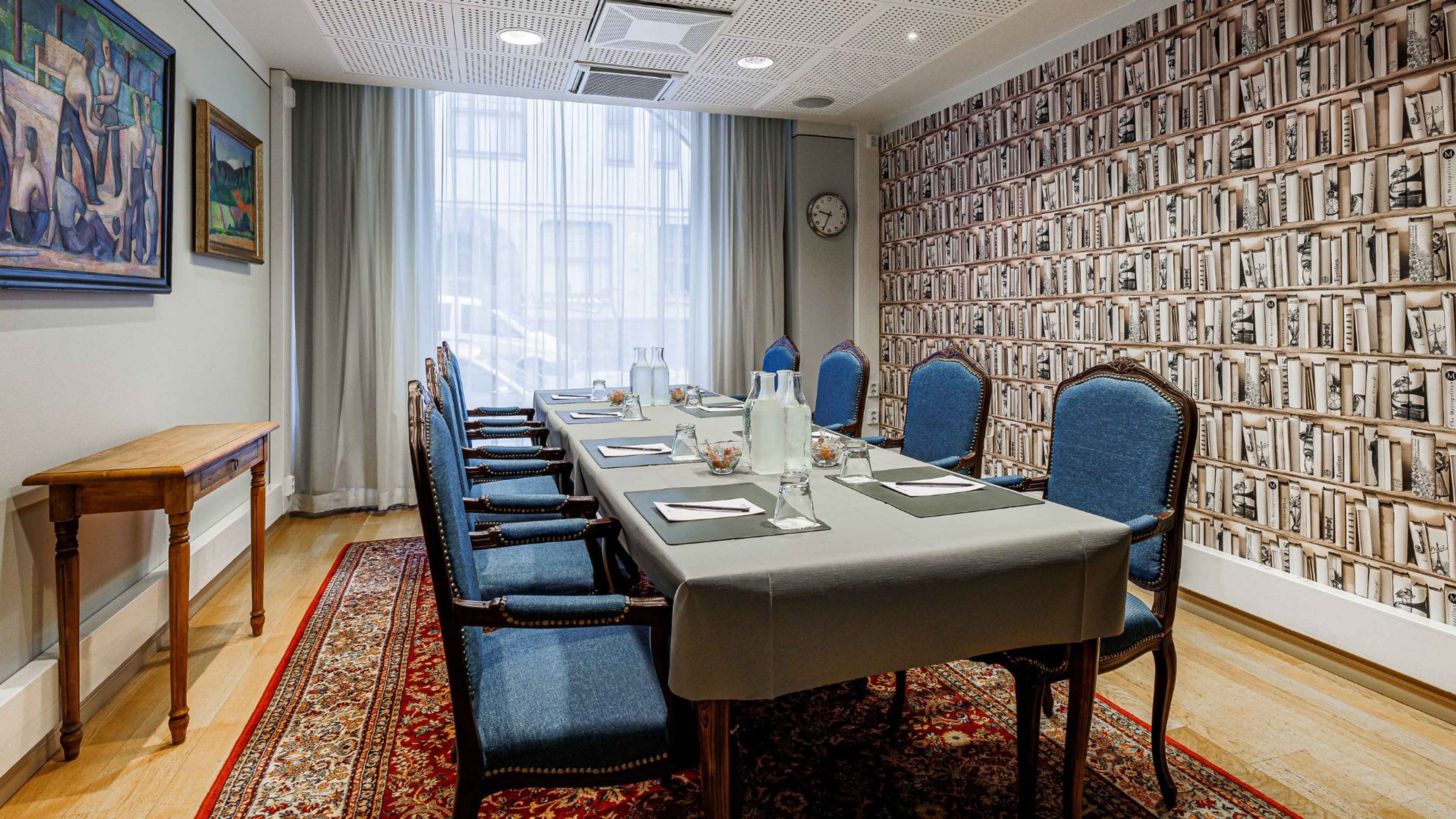 Kokoushuone kirjahylly-tapettiseinällä, pitkä pöytä sinisillä tuoleilla, puulattia, ikkunat ja taideteoksia seinällä.