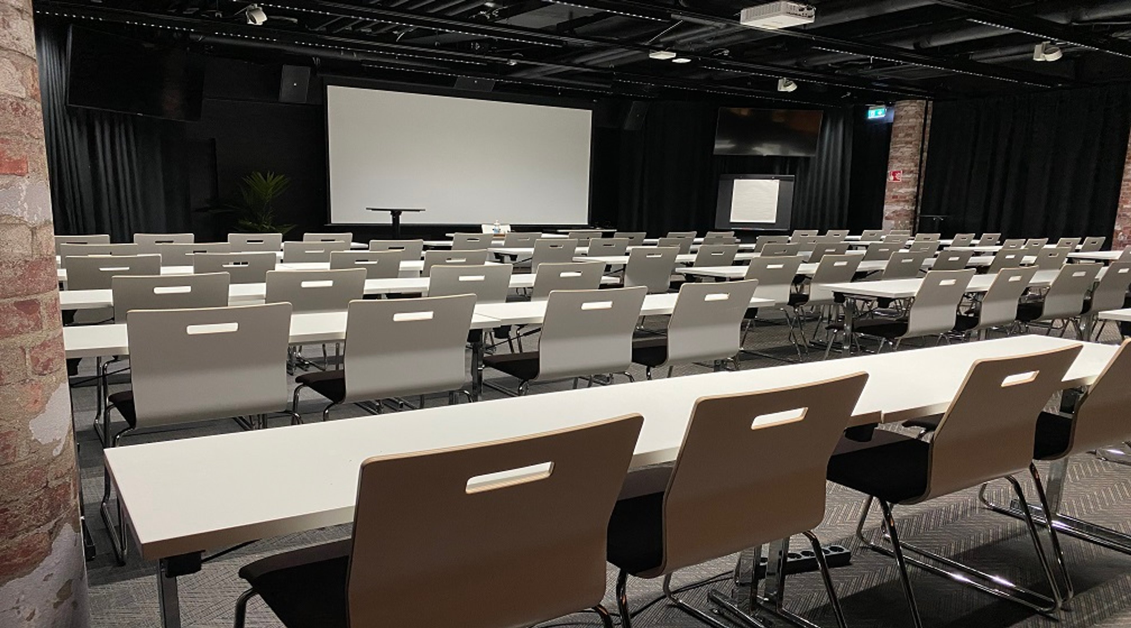 Auditorio valkoisten pöytien ja tuolien kanssa, valkokangas edessä ja tummilla verhoilla reunustettu. Tasainen kokolattiamatto lattialla.