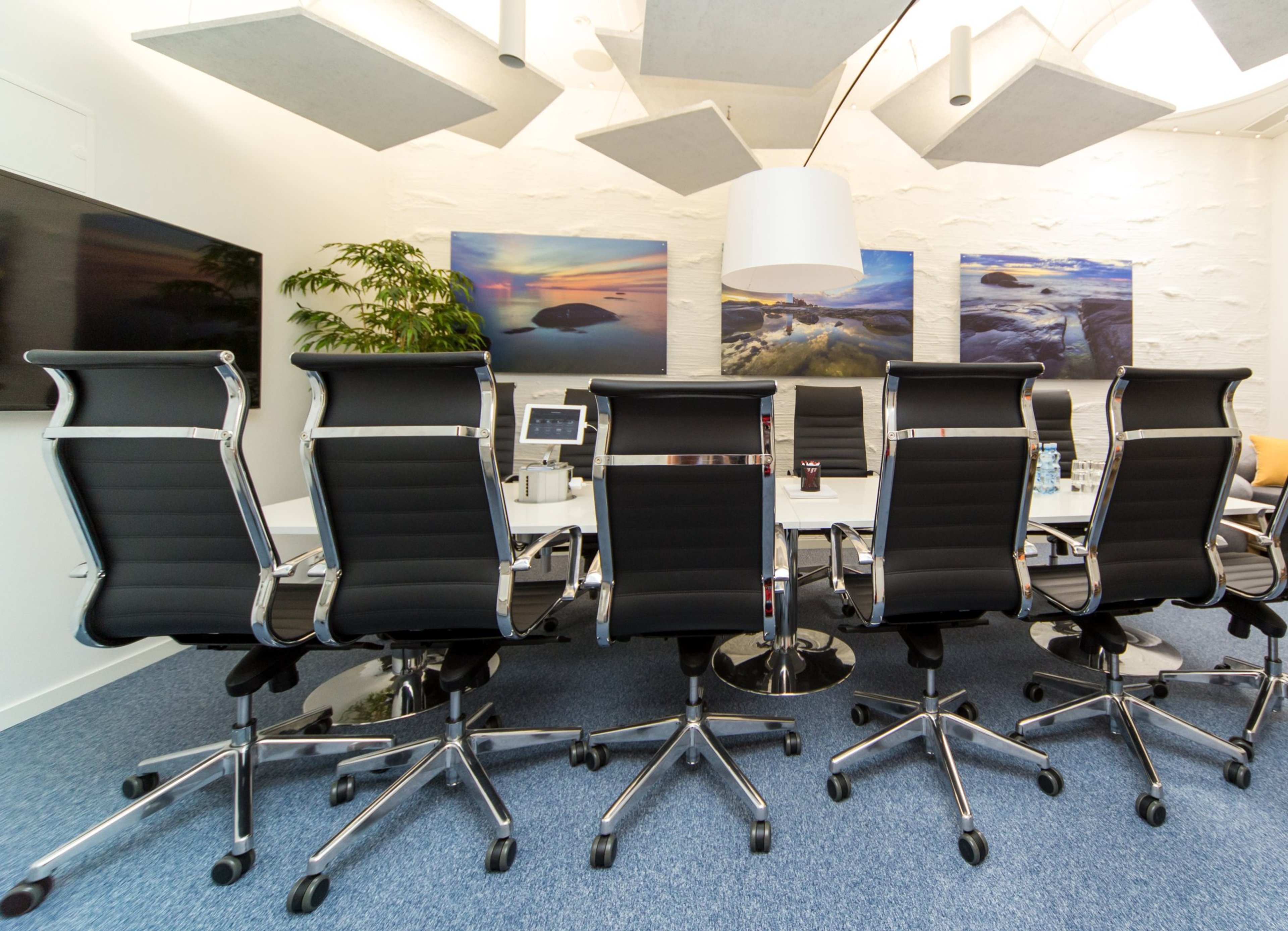 Moderni neuvotteluhuone sinisellä matolla, mustilla toimistotuoleilla, valkoisella pöydällä ja seinällä merimaisematauluja.