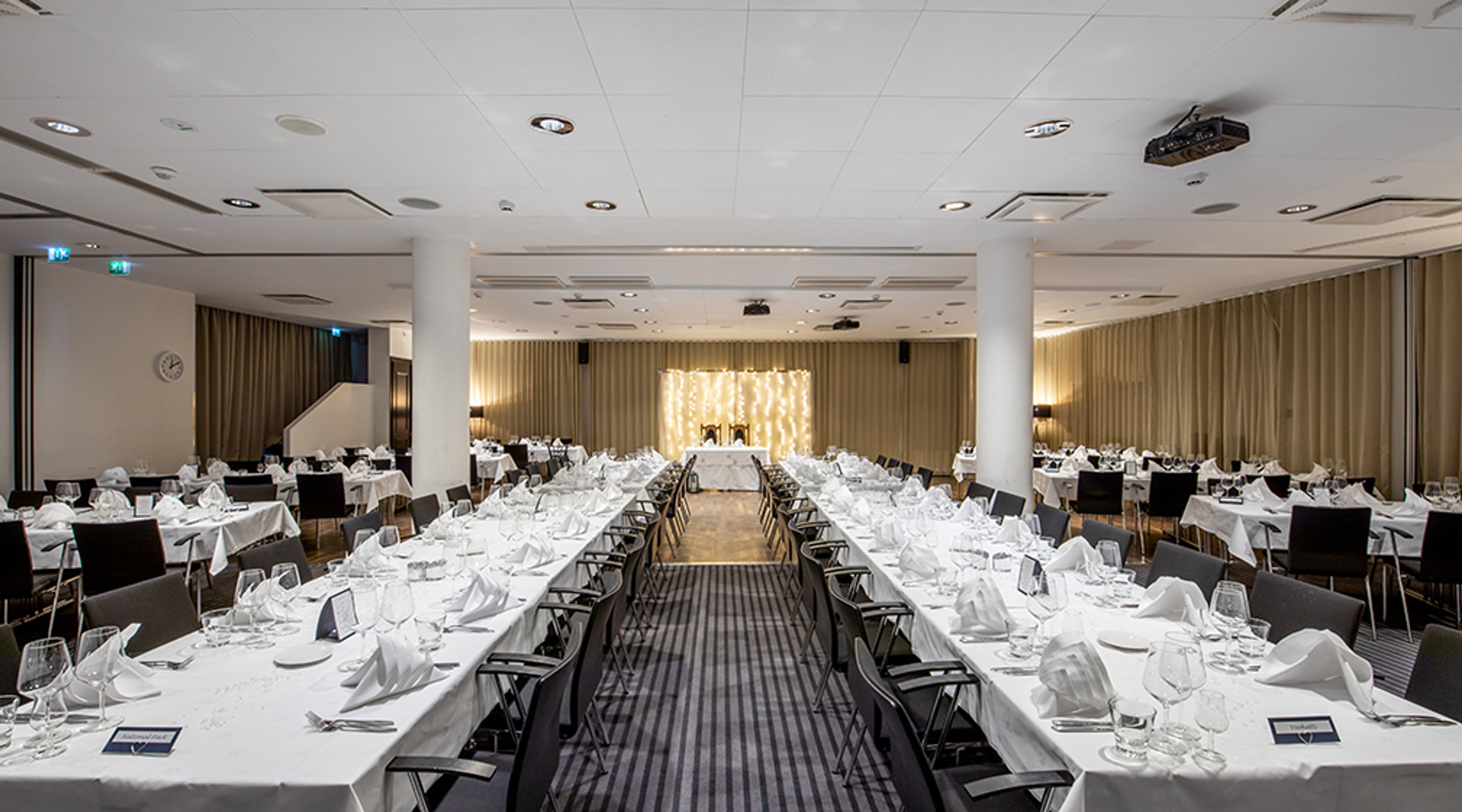 Juhlava ruokasali, jossa pitkät pöydät on katettu valkoisilla pöytäliinoilla ja taustalla on valaistu esiintymisalue. Tilassa isot pylväät.