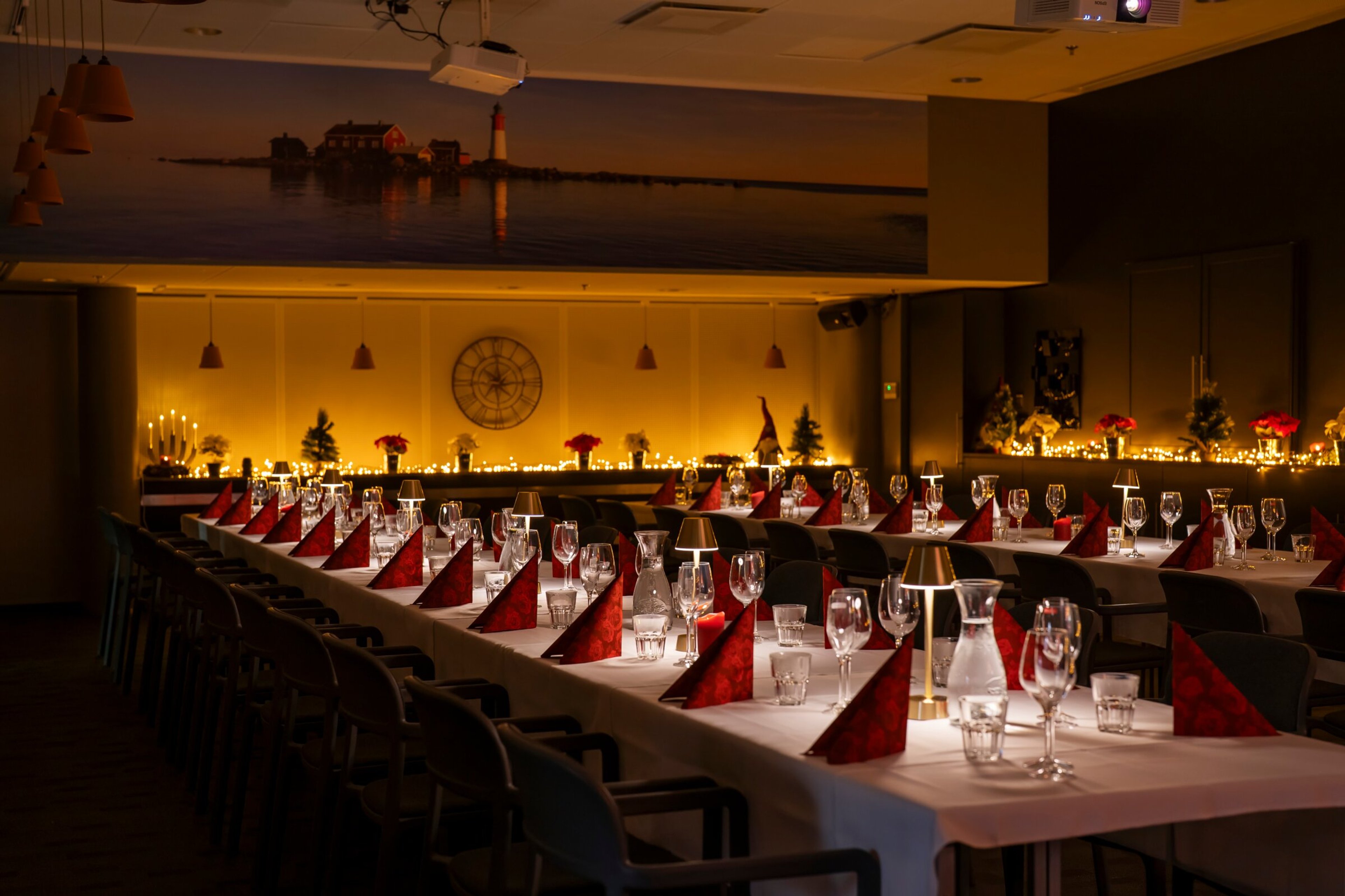 Ravintolan juhlava kattaus punaisilla lautasliinoilla, kynttilöillä ja lasiastiastoilla. Seinällä merimaisemataulu ja kello lämpimässä valaistuksessa.