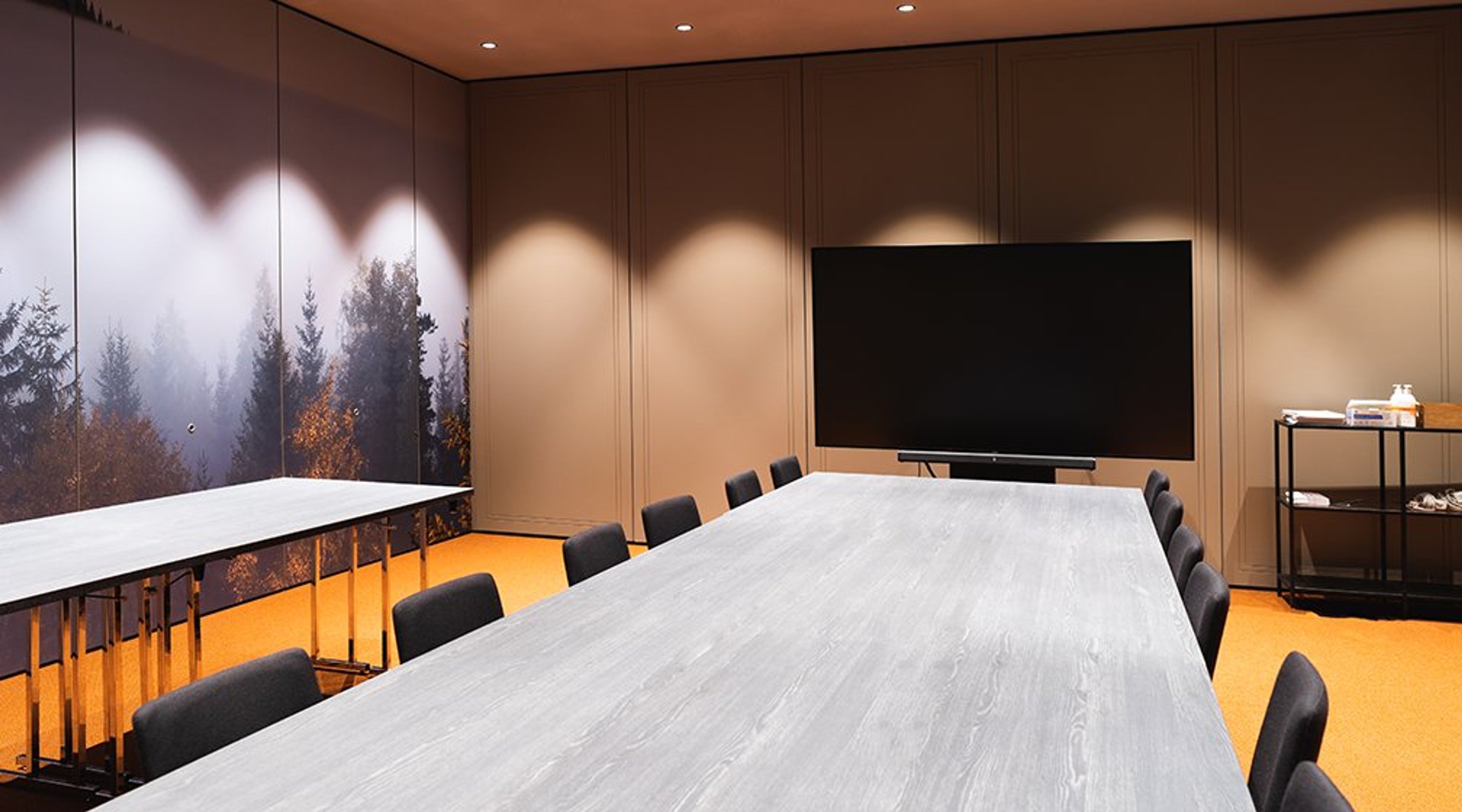 Moderni neuvotteluhuone, jossa vaalea puupöytä, mustat tuolit, seinällä metsäaiheinen kuvatapetti ja iso näyttö.