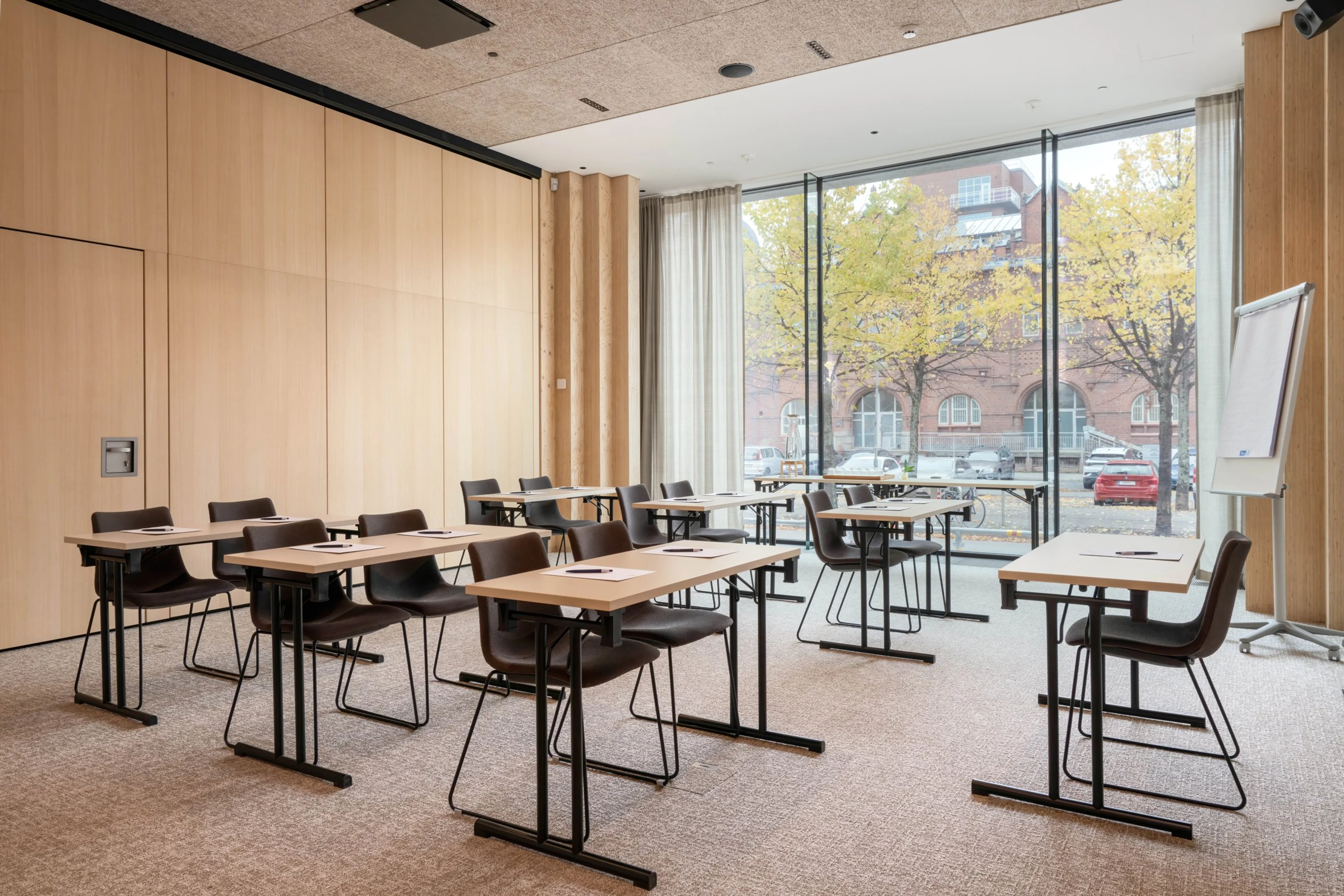 Modernt mötesrum med träpaneler, lätta bord och stolar. Rummet har heltäckningsmatta och stora fönster med utsikt mot en höstgata.