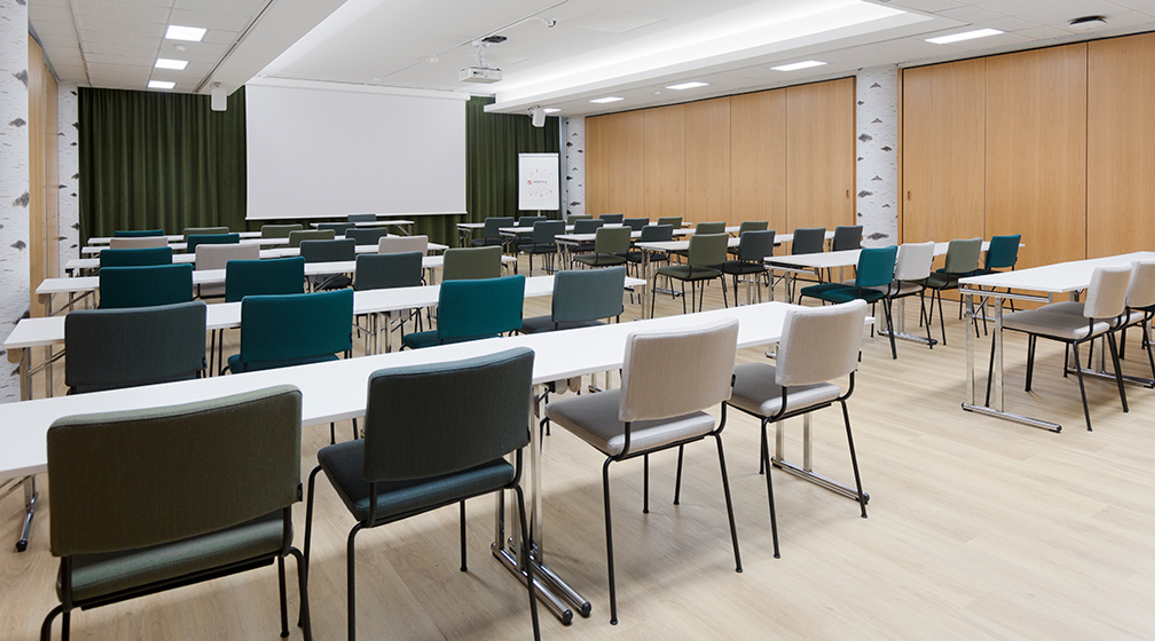 Valoisa kokoustila pitkillä pöydillä ja värikkäillä tuoleilla. Edessä valkokangas ja vihreät verhot. Taustalla puupanelointi ja tasainen puulattia.