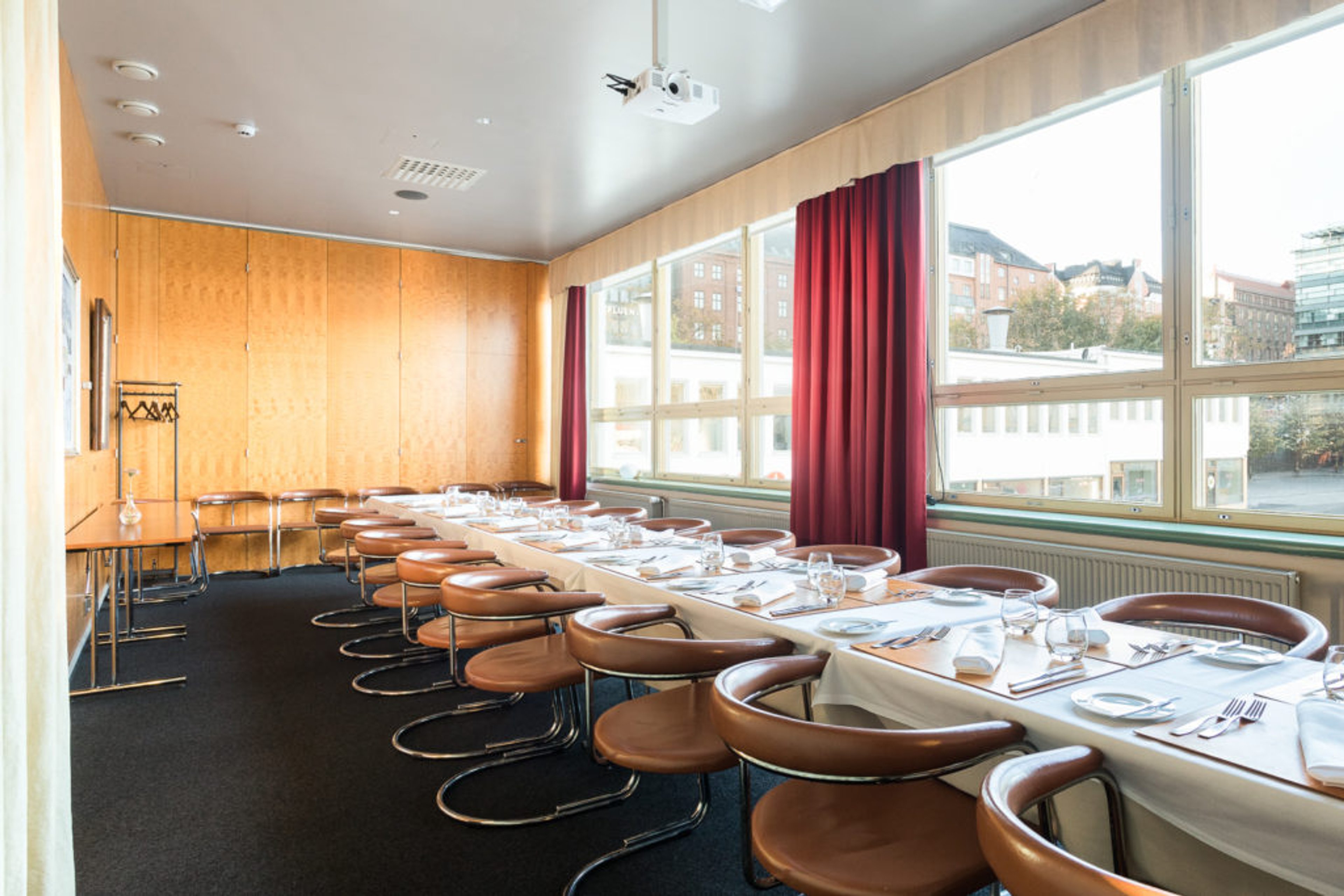 Mötesrum med träpaneler och långt bord med bruna stolar. Stora fönster och röda gardiner. Projektor hänger från taket.