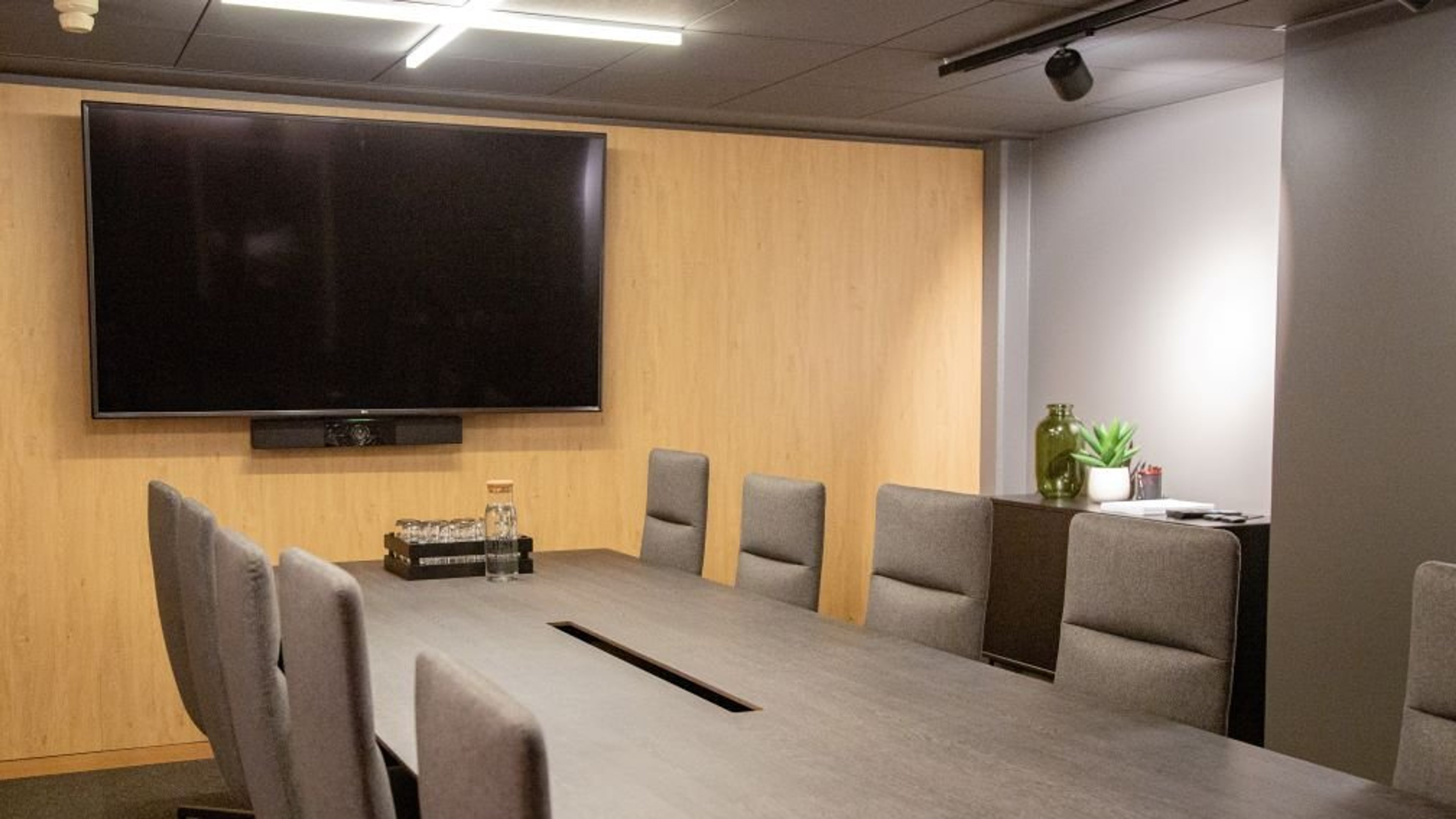 Moderni neuvotteluhuone, jossa suuri pöytä harmailla tuoleilla, seinällä näyttö ja puupaneelit sekä viherkasveja hyllyllä.