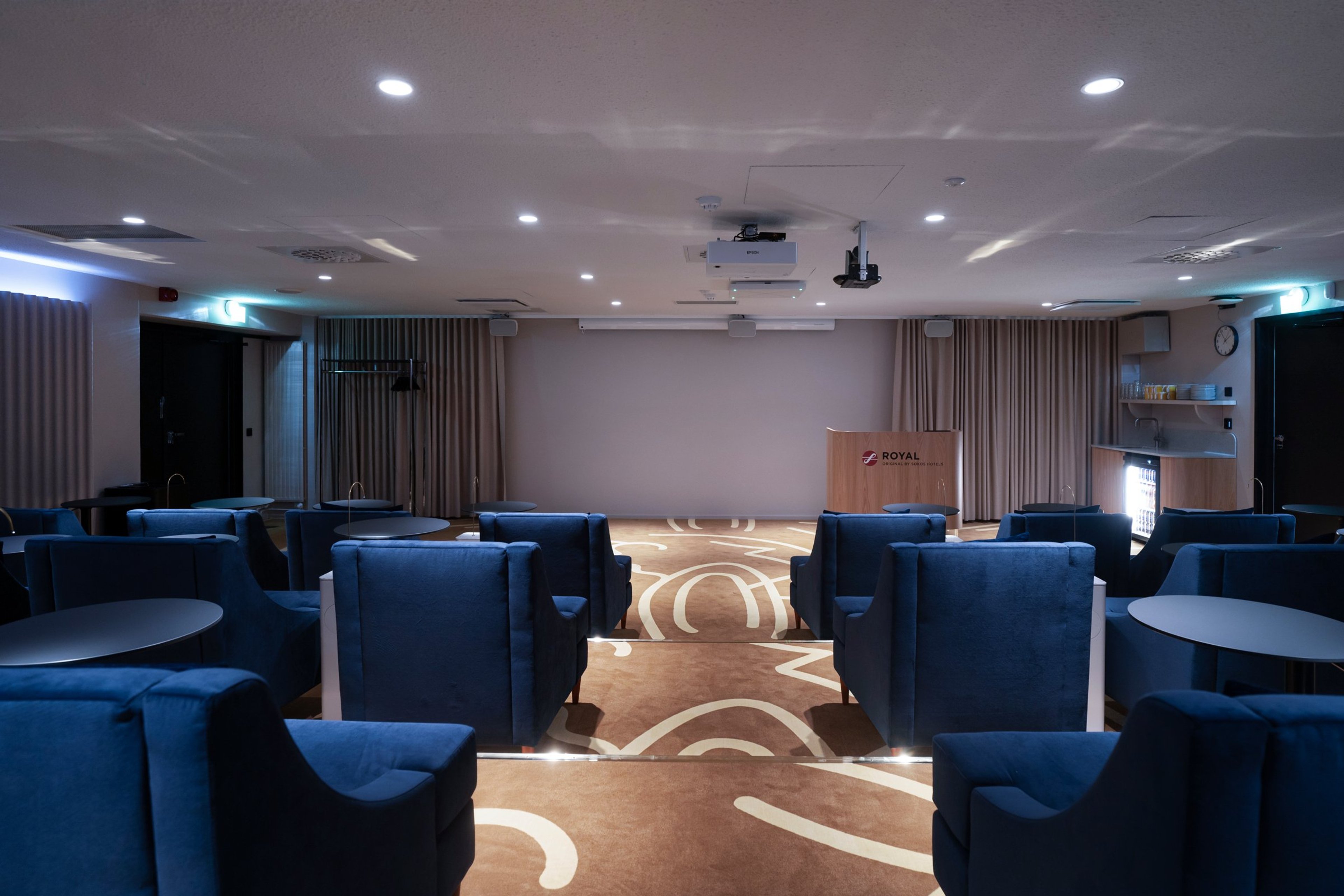 Konferenssihuone, jossa sinisiä nojatuoleja, puinen puhujanpönttö ja projektorit katossa. Tasainen kokolattiamatto, hyvä valaistus.