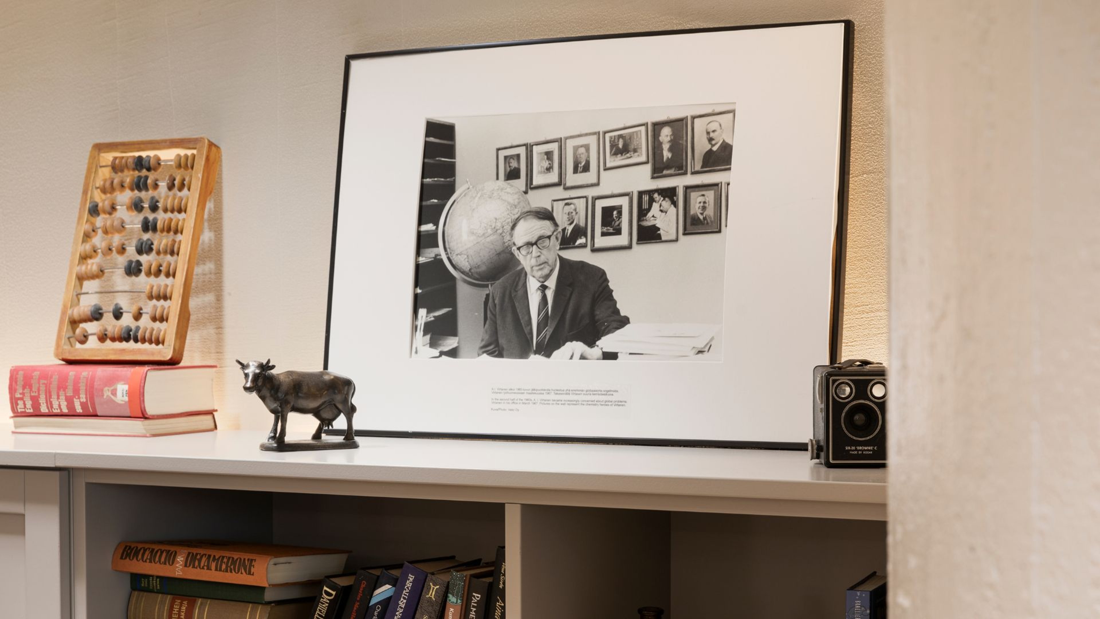 Valkoinen hylly, jossa historiallinen mustavalkoinen valokuva, helmitaulu, pieni lehmäpatsas ja vintage-kamera. Alla kirjahylly.
