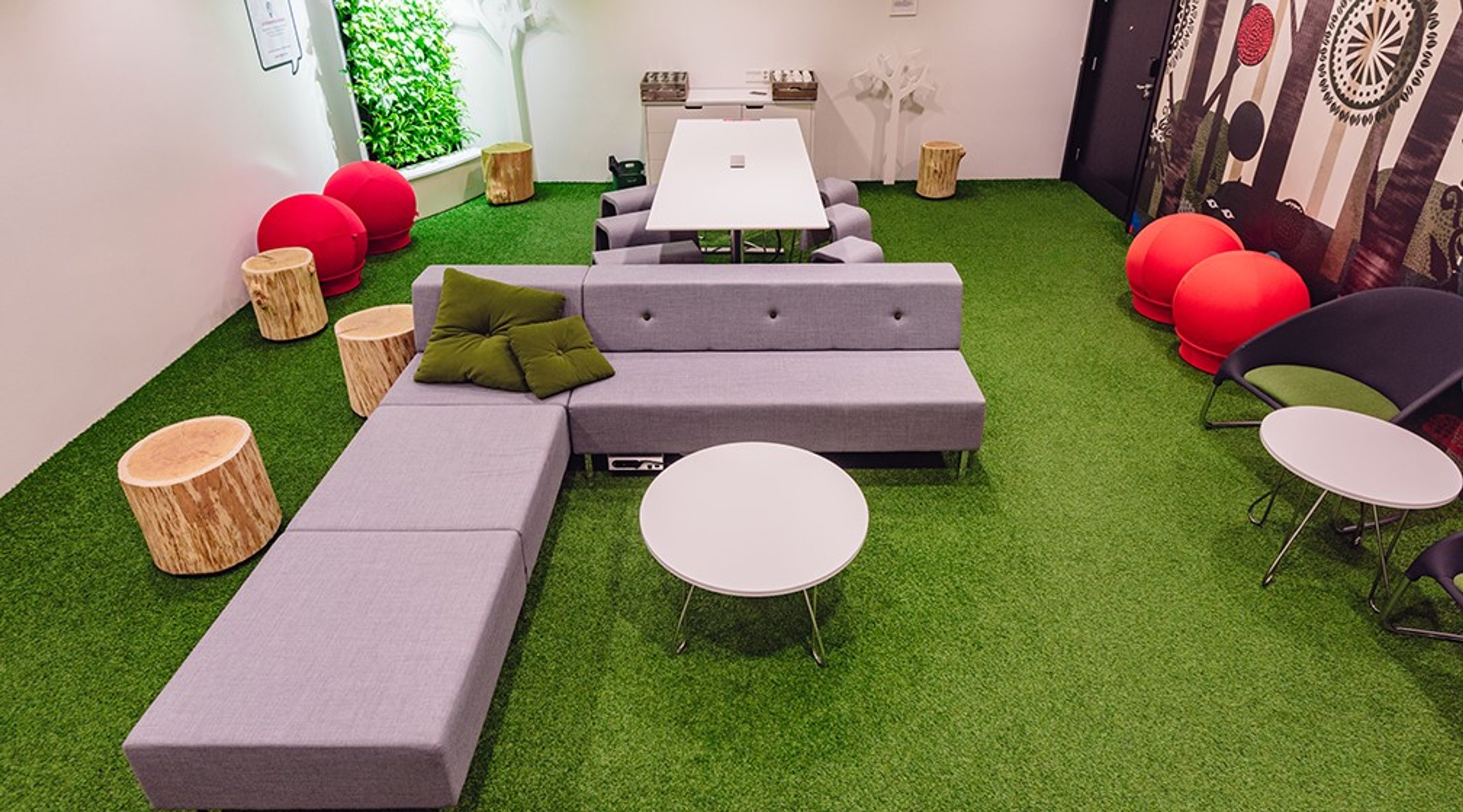 Leikkisä neuvottelutila, jossa vihreä nurmimattoalusta, harmaita sohvia, puisia pölkkyjä ja punaisia pallotuoleja. Valkoinen pöytä keskellä.