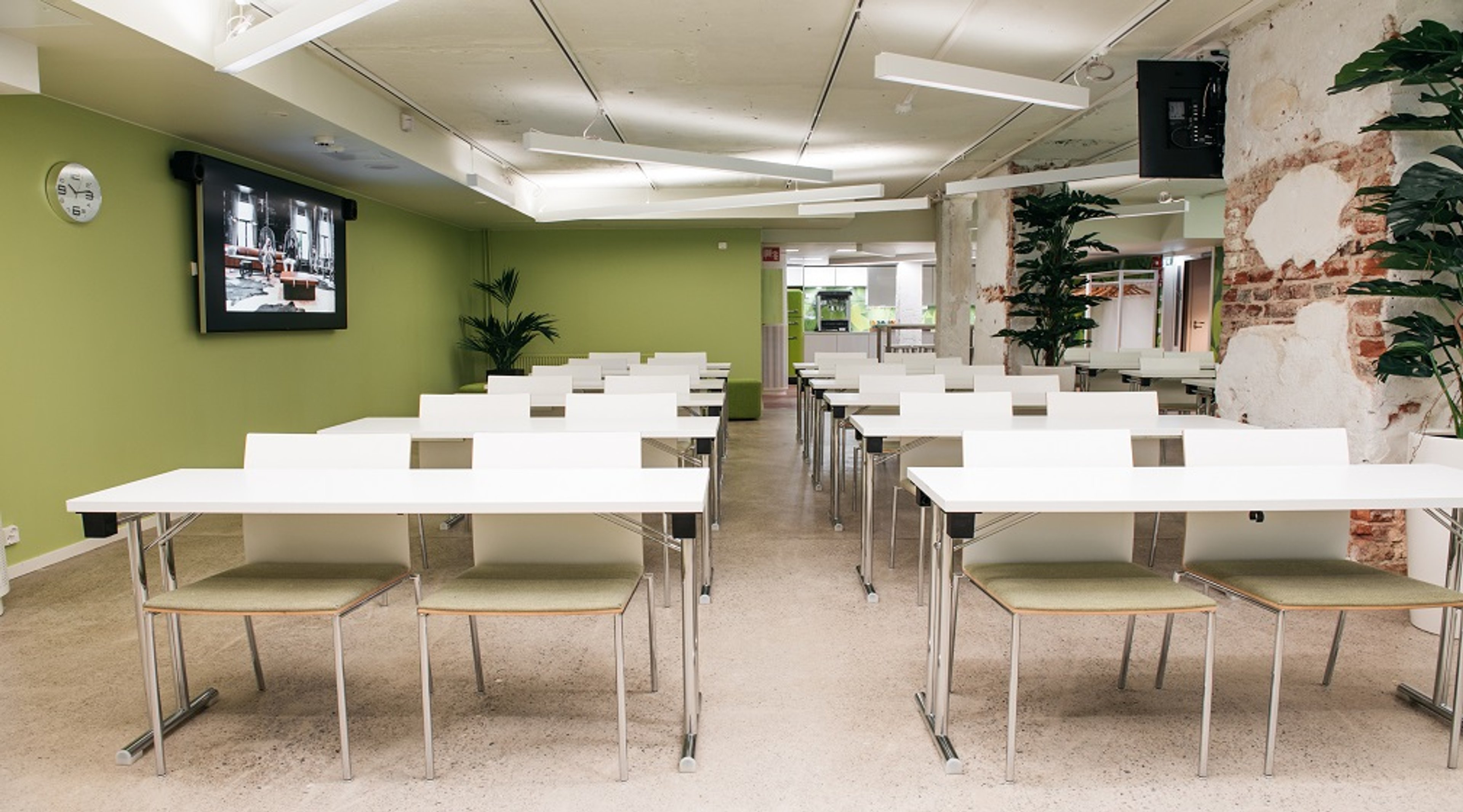 Kokoushuone valkoisilla pöydillä ja tuoleilla, vihreällä seinällä ja tiiliseinärakenteella. Näyttö seinällä ja viherkasveja huoneessa.