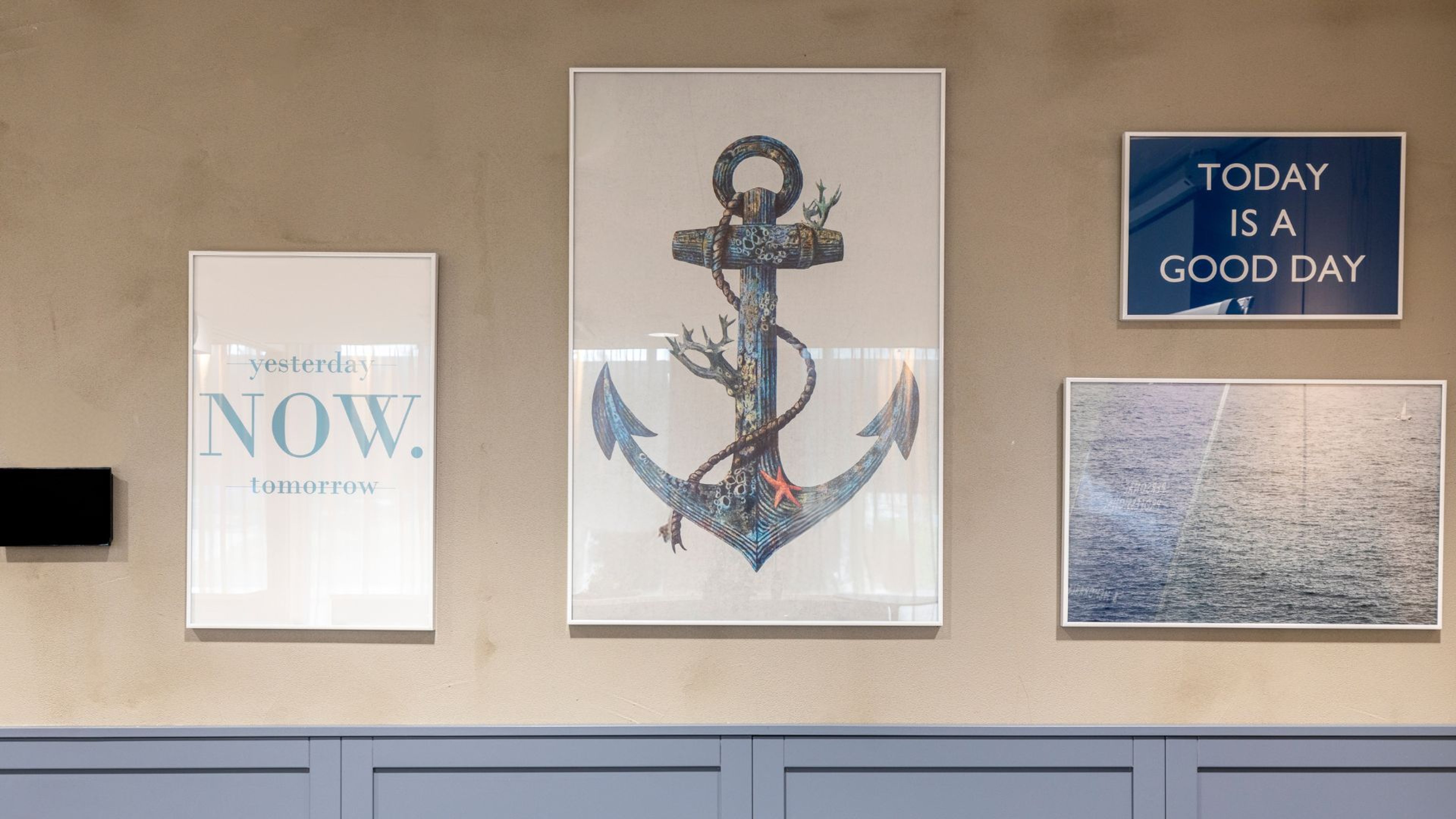 Neljä kehystettyä taulua vaalea-sävyisellä seinällä, keskellä ankkurikuvataulu, oikealla merimaisema.
