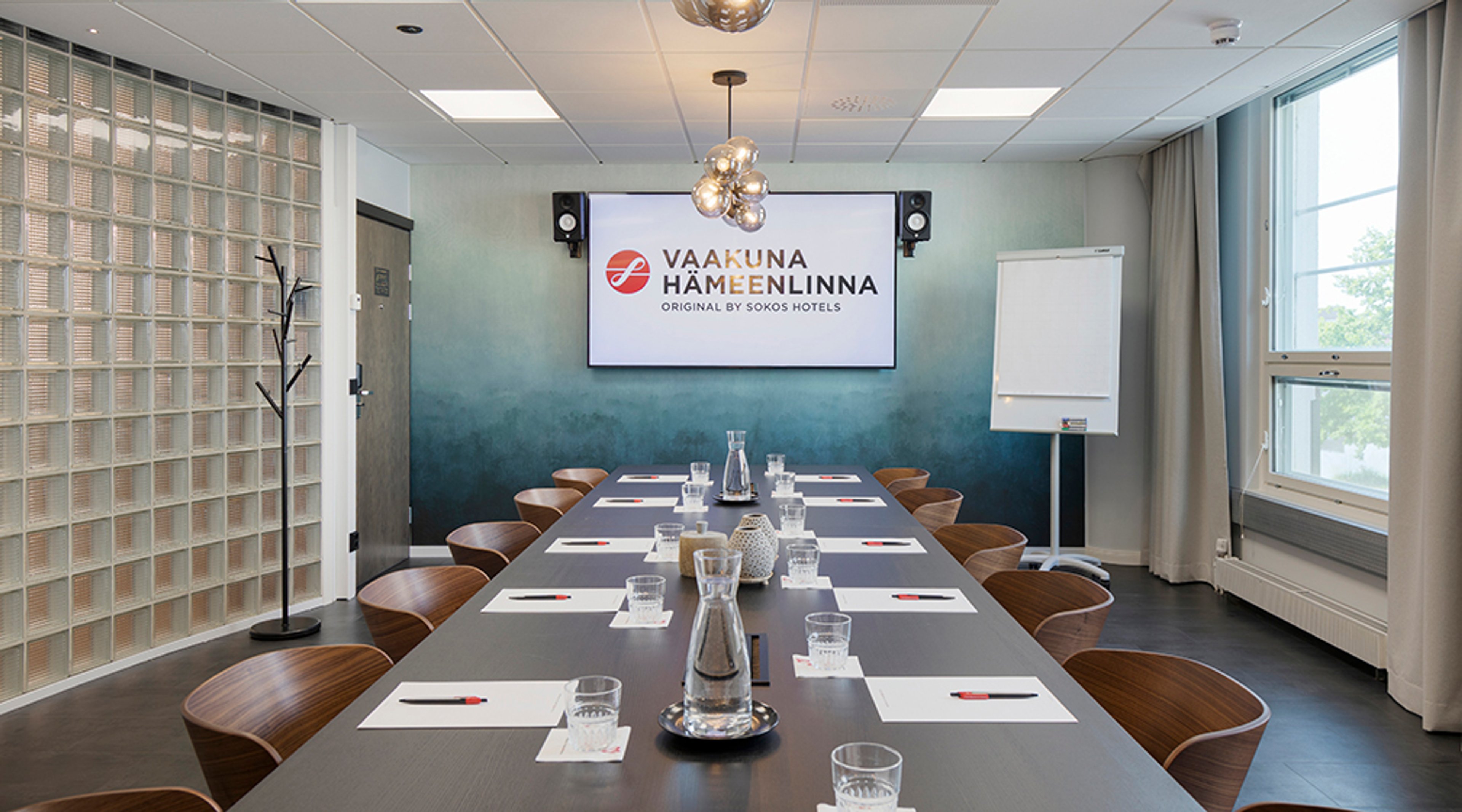 Kokoushuone pitkällä pöydällä, puiset tuolit, vesikarahvit, valkotaulu ja näyttö seinällä, sinisävyinen seinä ja isot ikkunat.