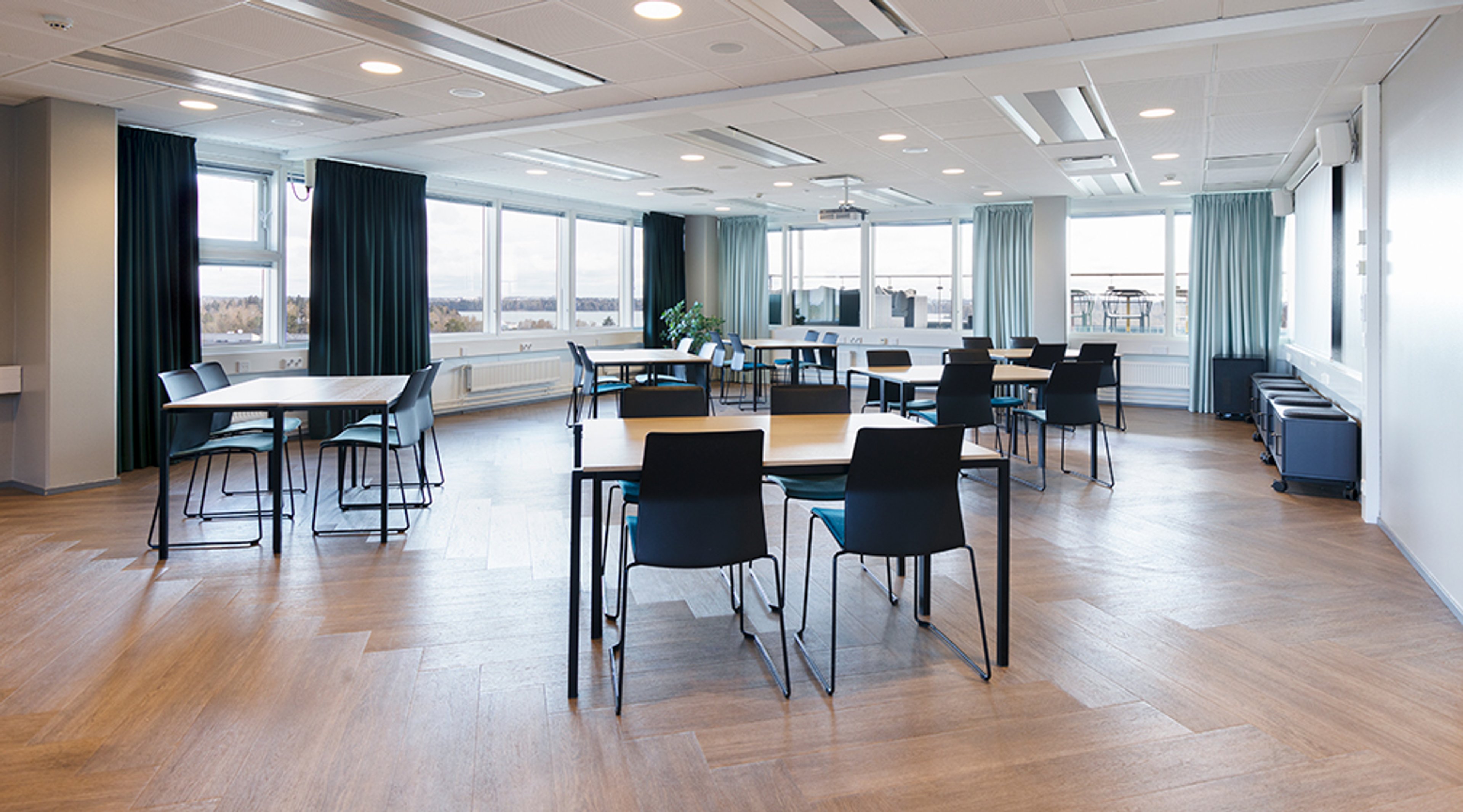 Bright meeting room with wooden flooring, multiple table arrangements and large windows with a view. Dark curtains and good overhead lighting.