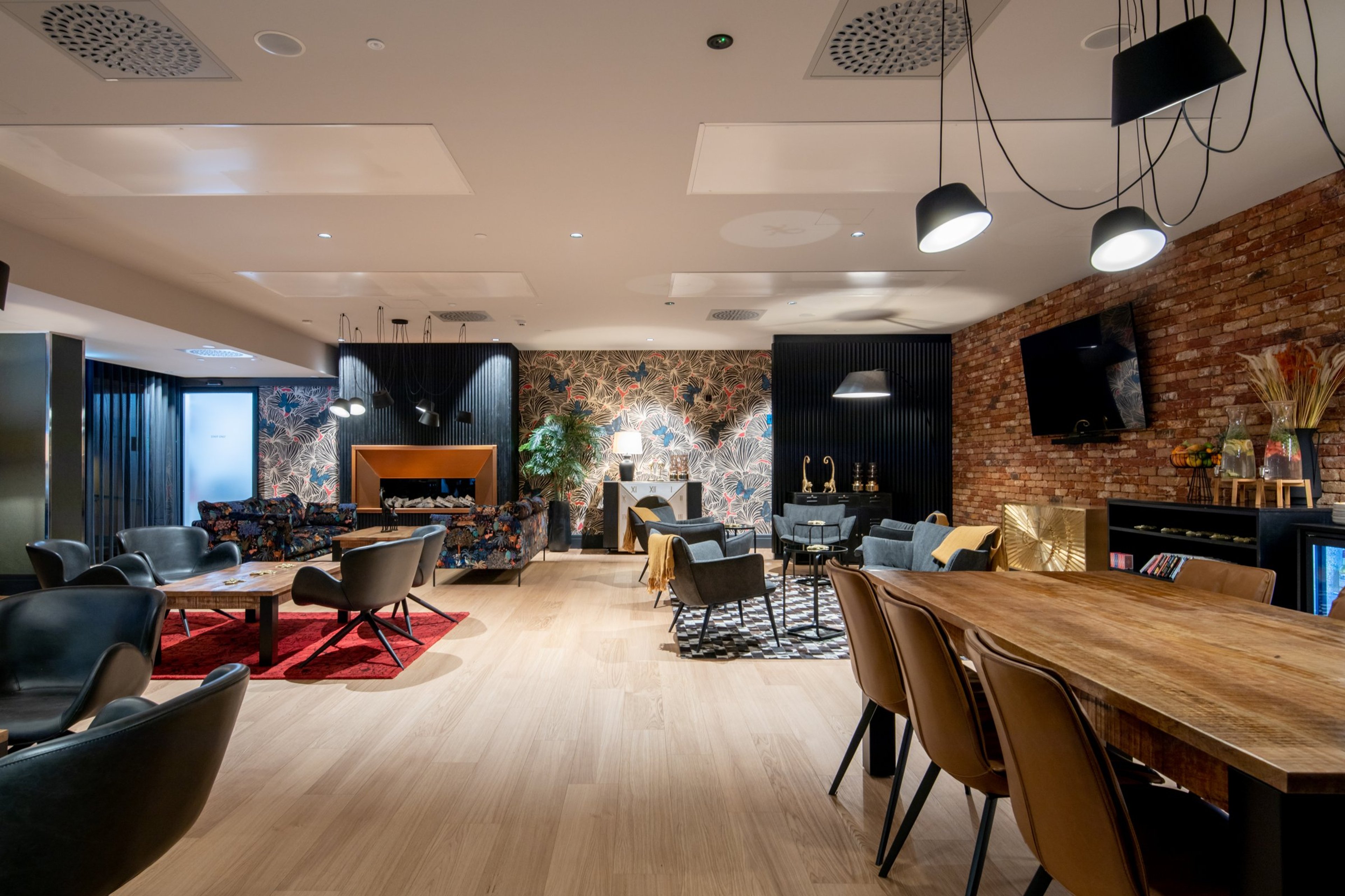 Moderni oleskelutila, jossa puulattia, tiiliseinä, koristeellinen tapetti, erilaisia istuinryhmiä ja puinen ruokapöytä. Tilassa on tasainen lattia ja hyvä valaistus.