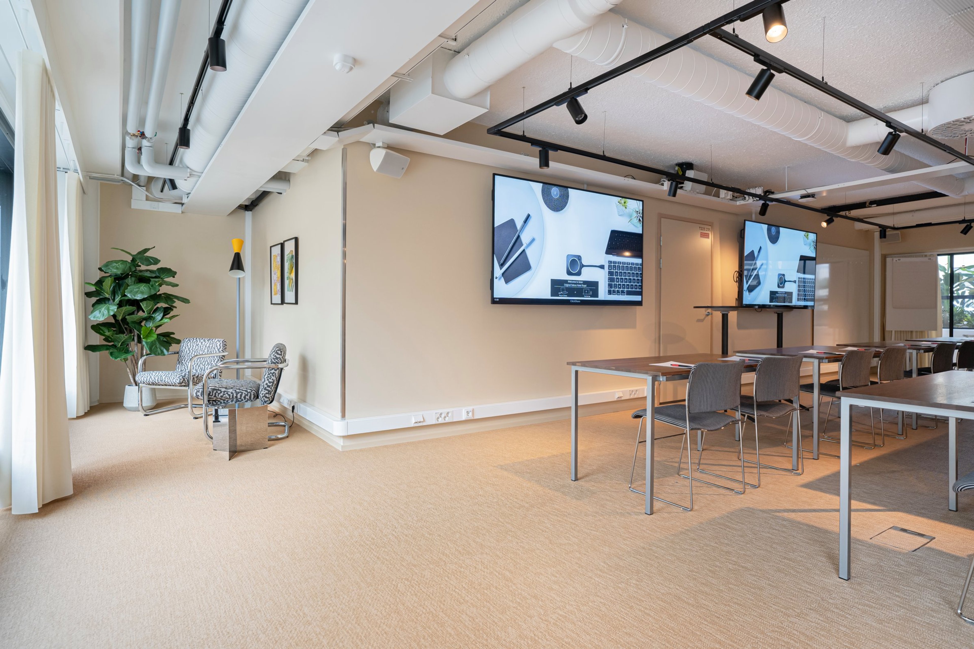 Moderni kokoustila beigen sävyisellä matolla, suurilla näytöillä, pöydillä ja tuoleilla. Tilassa on viherkasvi ja tyylikkäät valaisimet.